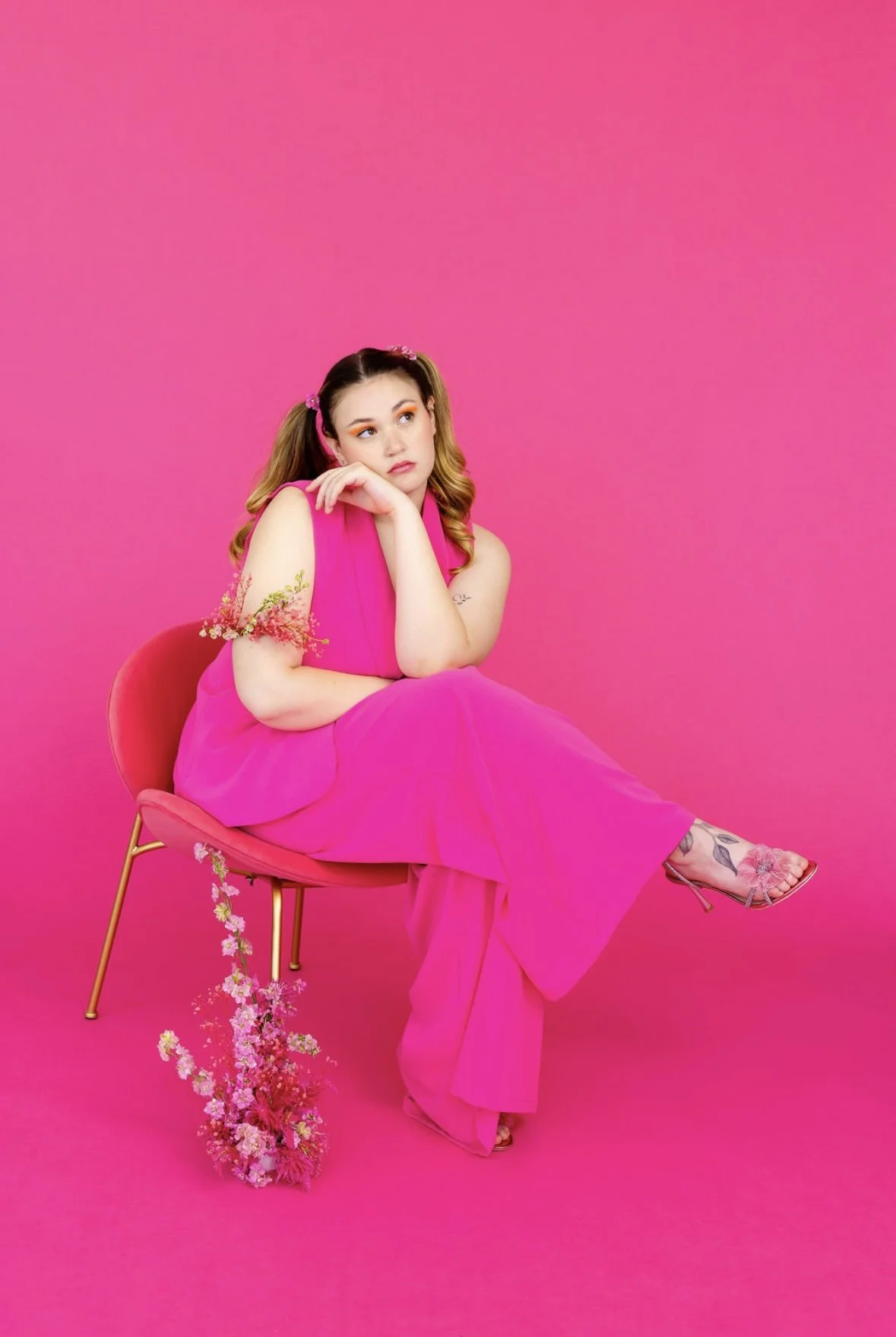 A young woman with light skin and wavy hair sits on a pink chair against a pink background, wearing a bright pink outfit and pink floral high-heeled shoes. She rests her chin on her hand, looking contemplative, with a small bouquet of pink and white 