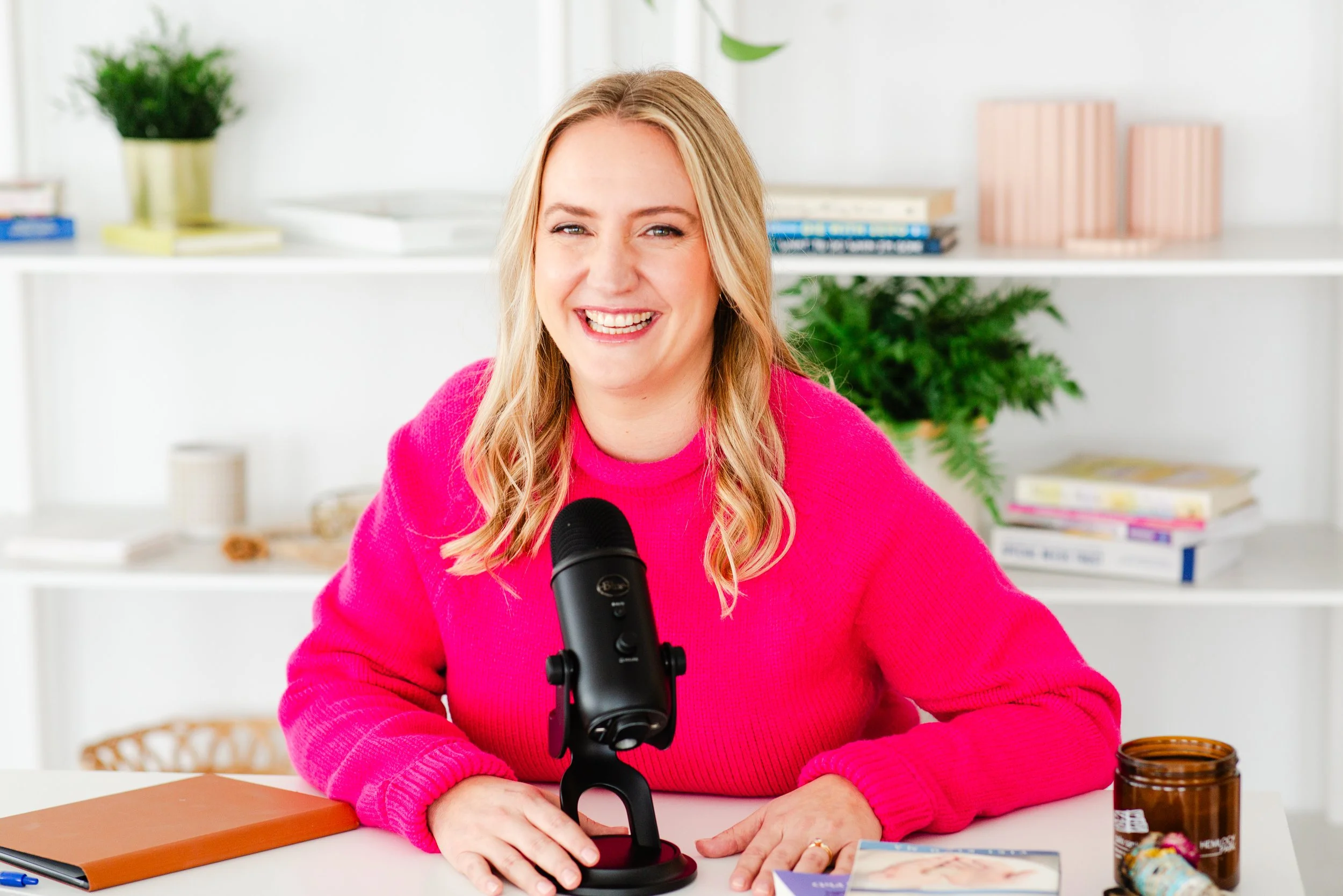 A woman in a bright pink sweater smiling and sitting at a desk with a microphone in front of her, with books and plants in the background.