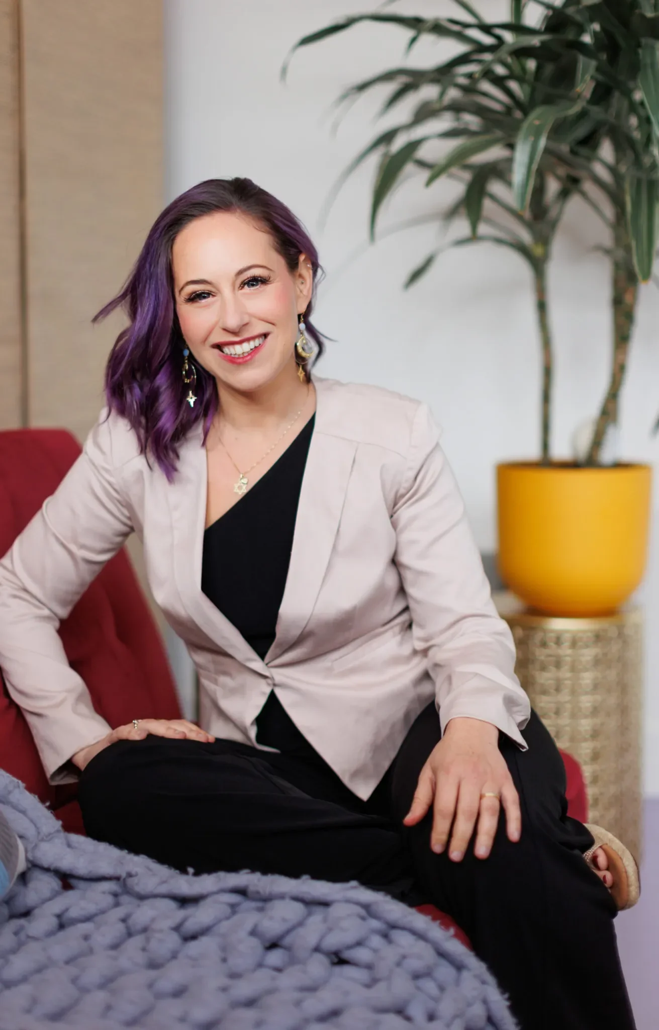 A woman with purple hair and earrings smiling at the camera, sitting on a red chair with a gray chunky knit blanket in front of her and a large potted plant in a yellow pot behind her.