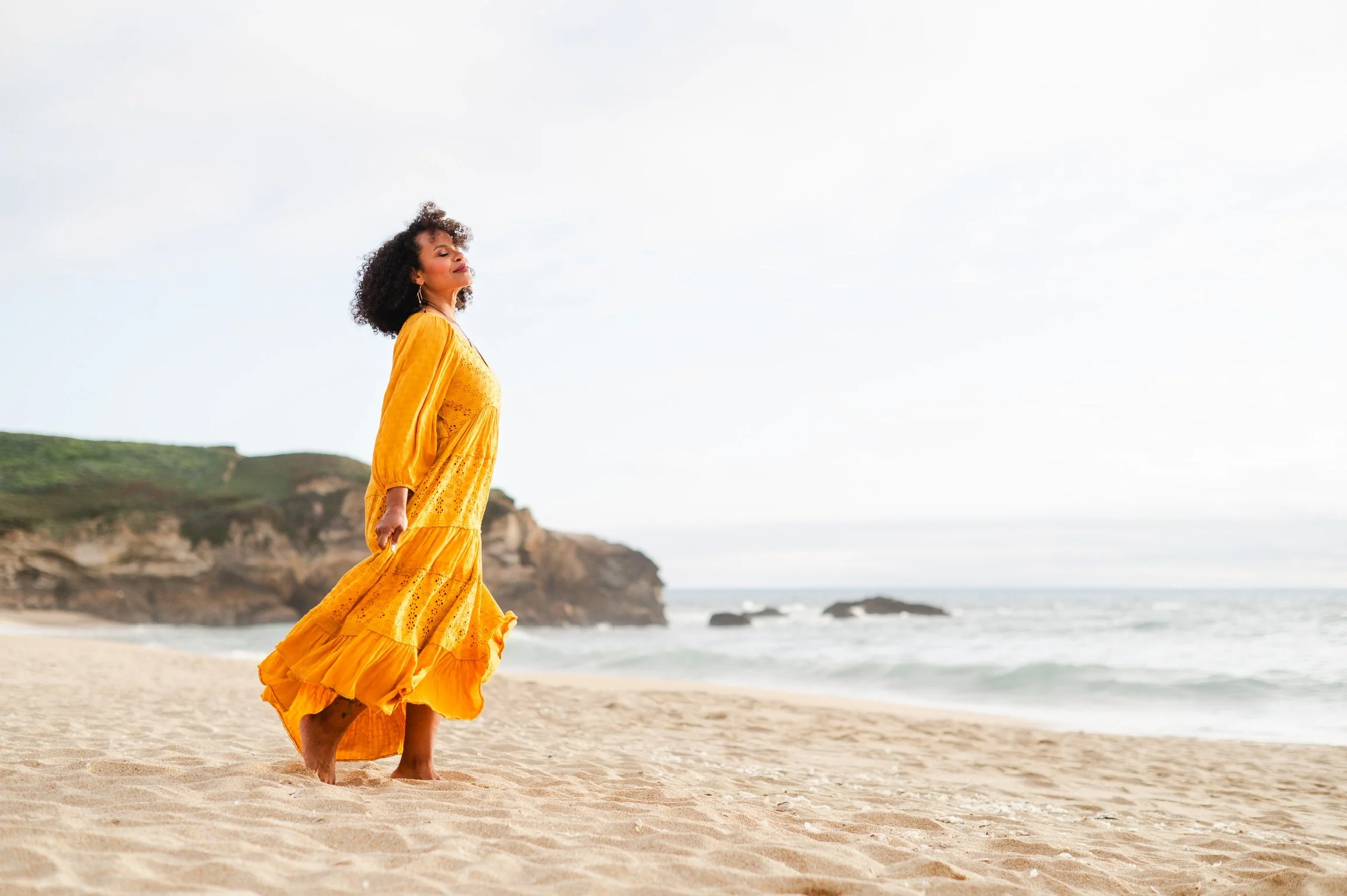 A woman in a yellow dress walking barefoot on a sandy beach with cliffs and the ocean in the background.