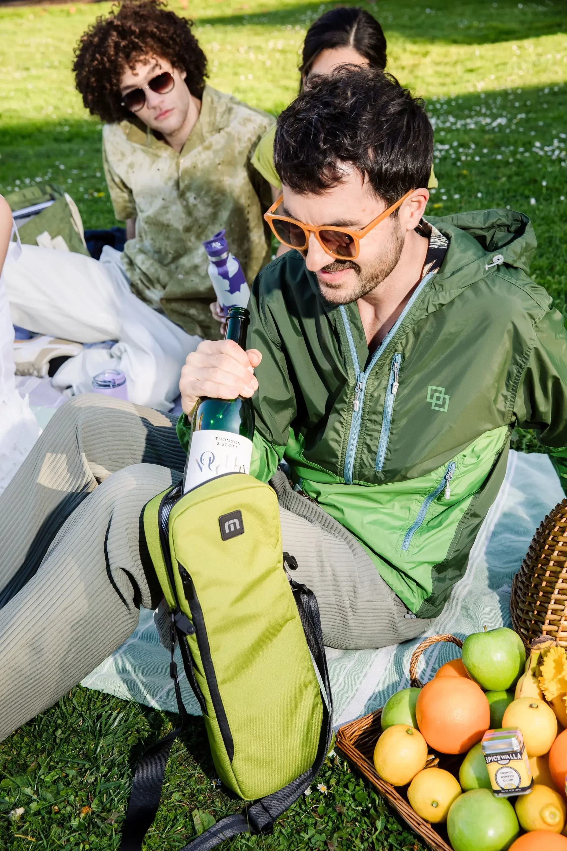 Group of friends enjoying a picnic outdoors, with one person holding a bottle, surrounded by apples, oranges, and snacks on a blanket.