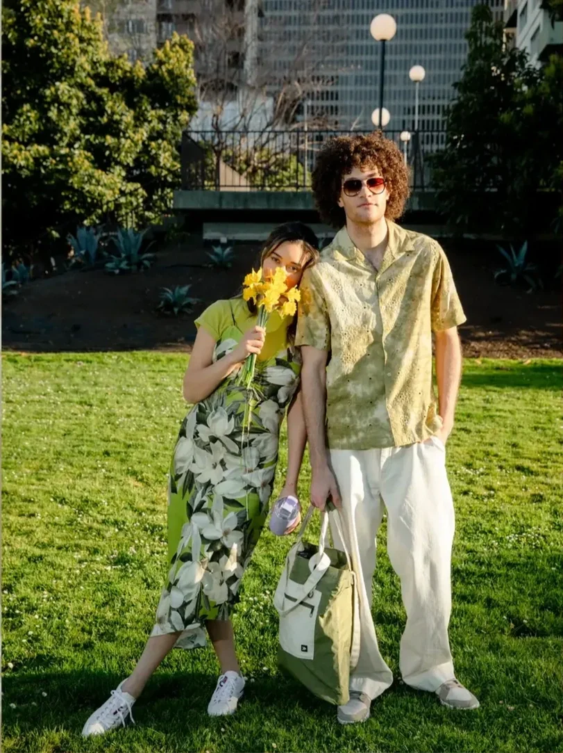 A young woman and man standing on a grassy park. The woman holds a bouquet of yellow flowers and leans her head on the man's shoulder. The man has curly hair, sunglasses, a yellow patterned shirt, and white pants, holding a reusable bag and a water b