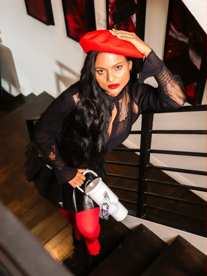 A woman with long black hair, red lipstick, and a black lace blouse, leaning on a staircase railing while holding a white water bottle. She is wearing a red beret, red tights, and black boots. The background includes framed artwork on the wall.