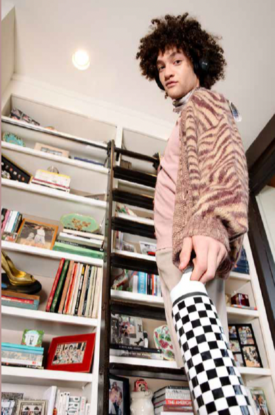 A young person with curly hair wears headphones, a pink shirt, a patterned cardigan, and checkered pants, standing indoors in front of a bookshelf filled with books and decorative items.