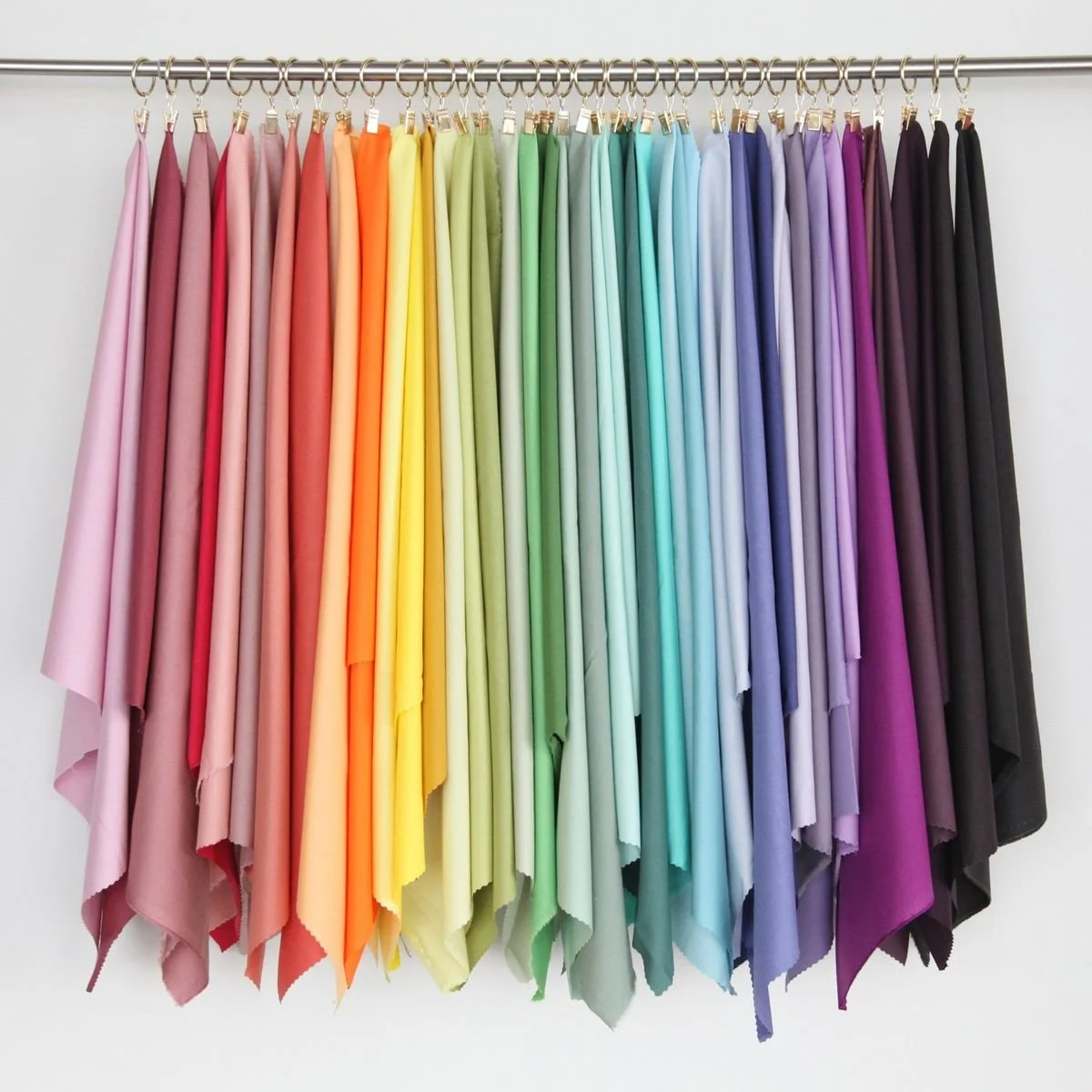 A rainbow of fabric pieces hanging on a metal rod, arranged from pink to black in color.
