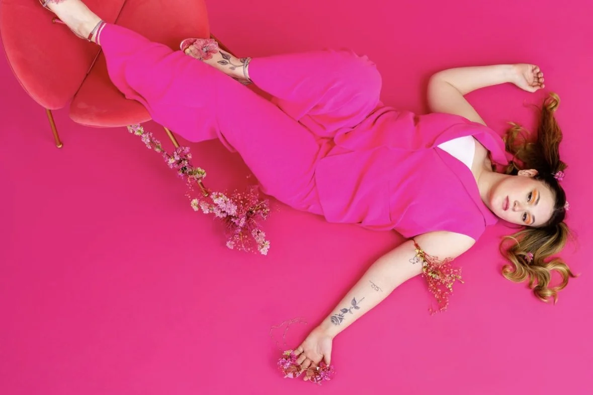 A woman with long, wavy hair lying on a pink background, wearing a pink dress and surrounded by pink flowers and a small pink chair.