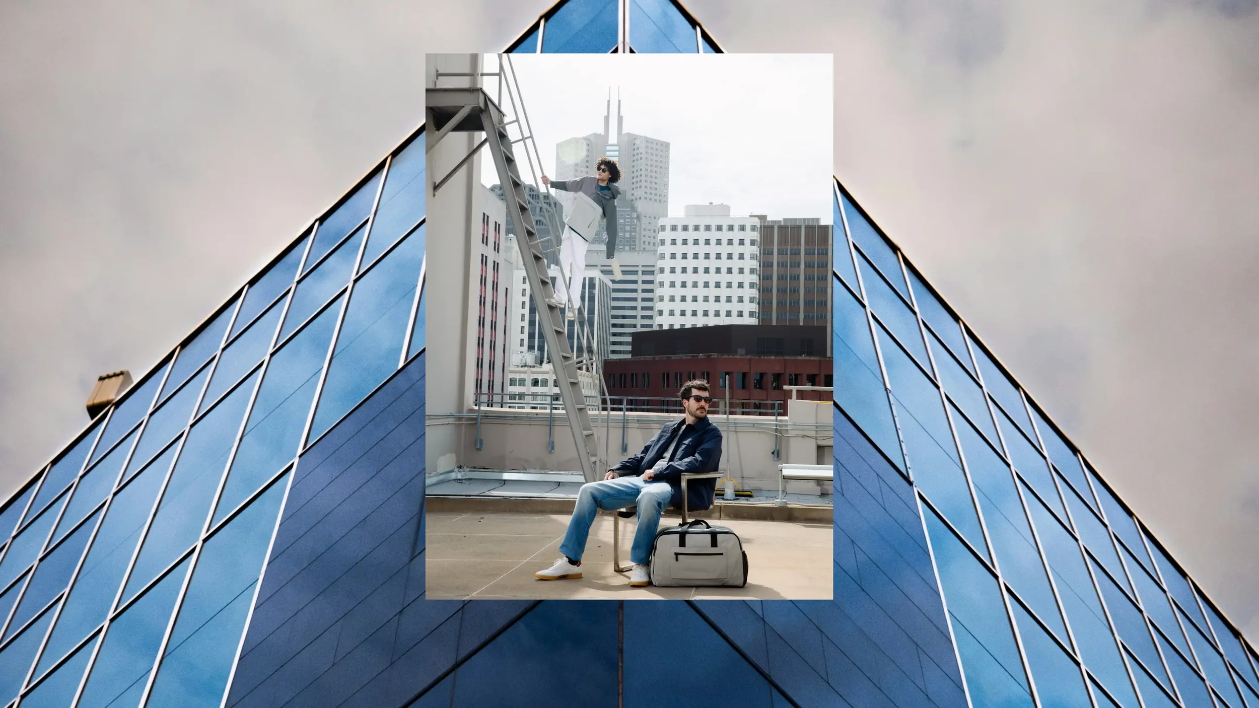 A man sitting on a chair on a rooftop with city skyscrapers in the background, wearing sunglasses, a dark jacket, jeans, and white sneakers, with a gray duffel bag beside him. Behind him, a woman in casual attire, including sunglasses, is climbing a 