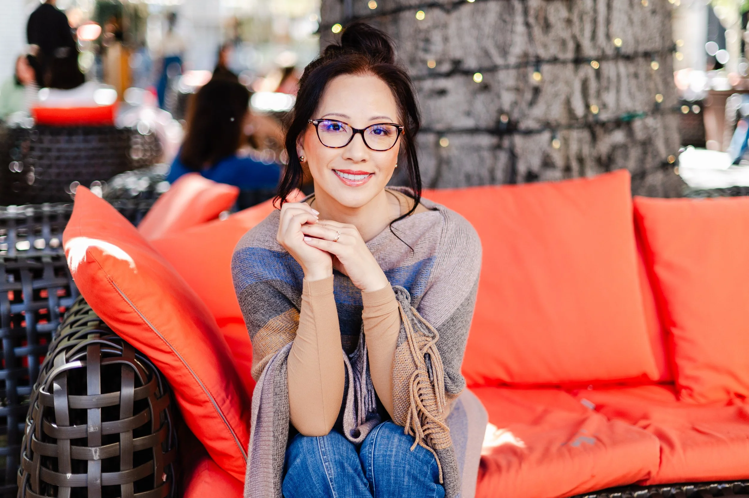 A woman with glasses and dark hair, smiling, sitting on a bright orange outdoor sofa with large cushions, in a patio setting.