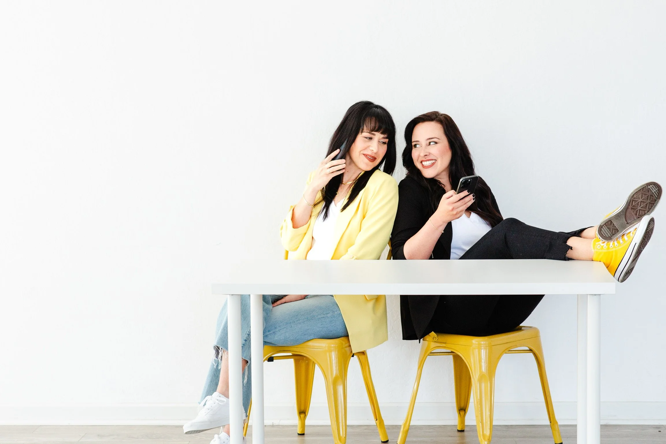 Two women sitting on yellow chairs behind a white table, smiling and looking at each other. One woman is wearing a yellow blazer and the other is wearing a black blazer. The woman in black is holding a smartphone and has her feet up on the table.