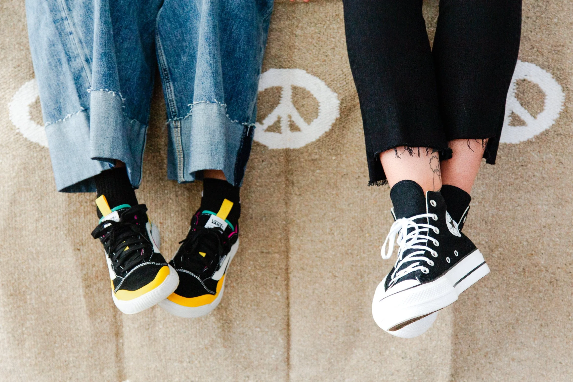 Two individuals sitting on a carpeted floor with peace symbols, wearing black and white sneakers, with one wearing ripped jeans and the other wearing frayed black pants.