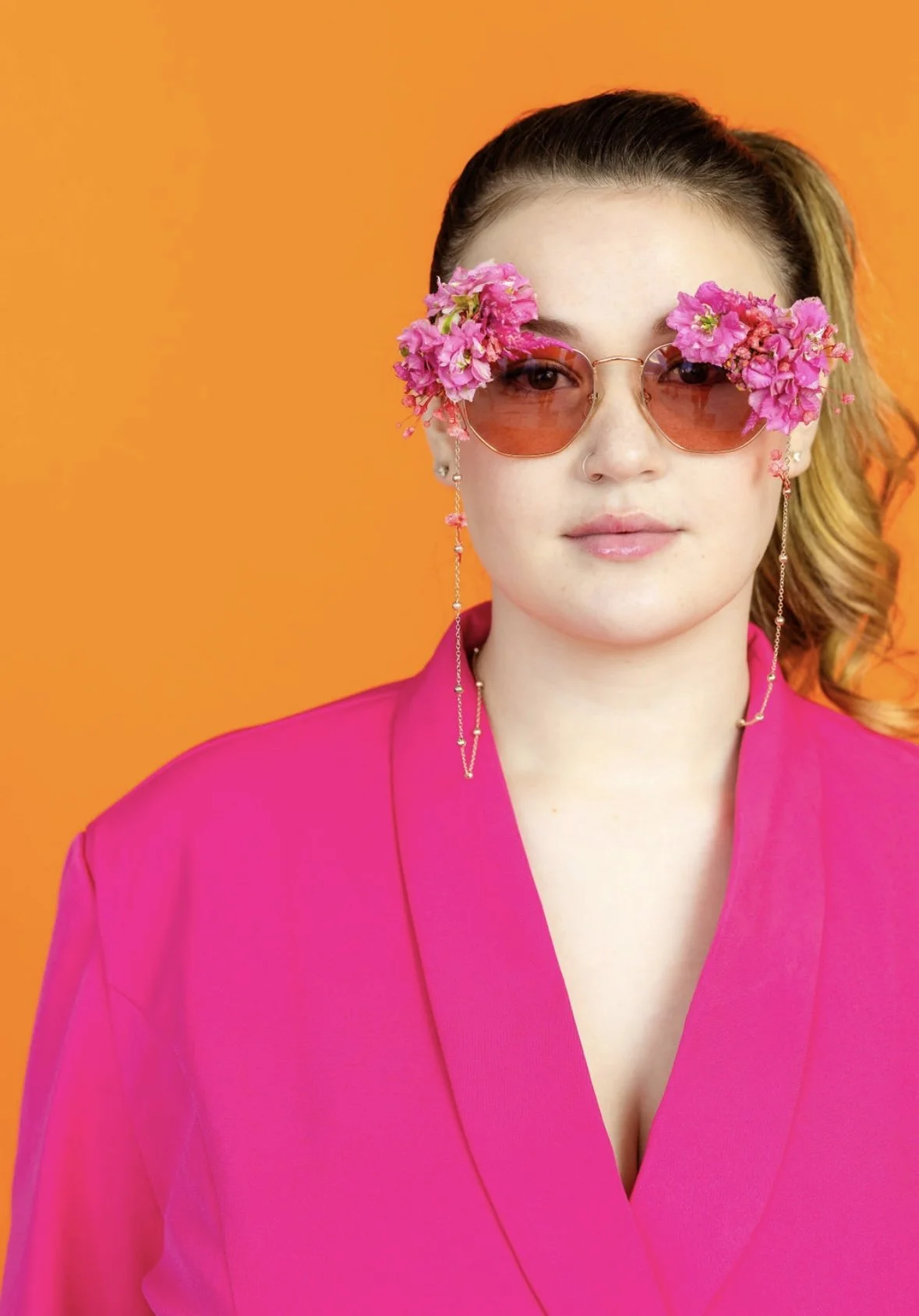 A woman wearing pink sunglasses decorated with pink flowers, a pink blazer, and long earrings, standing against an orange background.