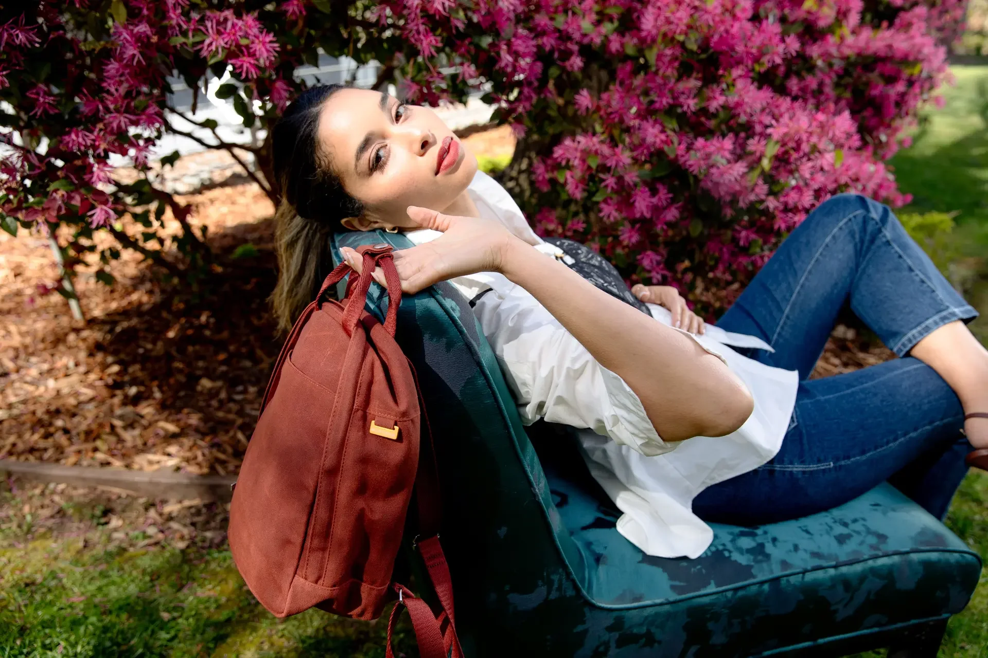 Woman reclining on a park bench with pink blooming bushes in the background, carrying a red backpack.