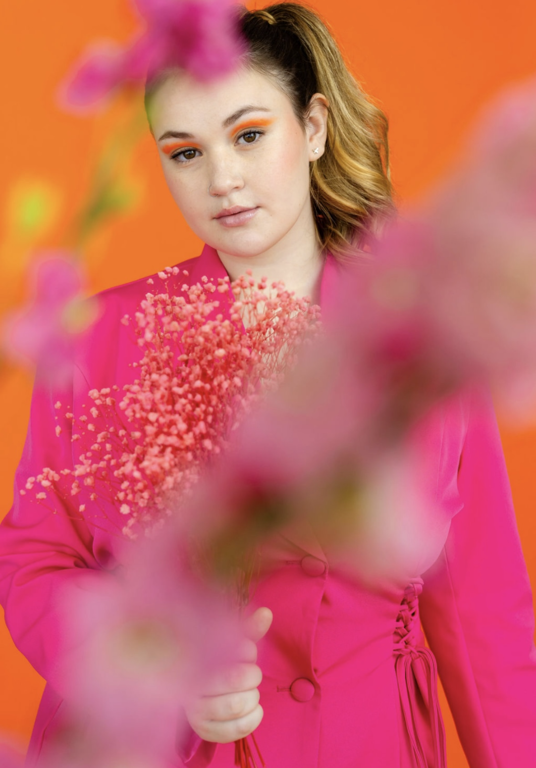 Young woman with shoulder-length wavy hair wearing a bright pink blazer and matching skirt, holding a pink bouquet, against an orange background.