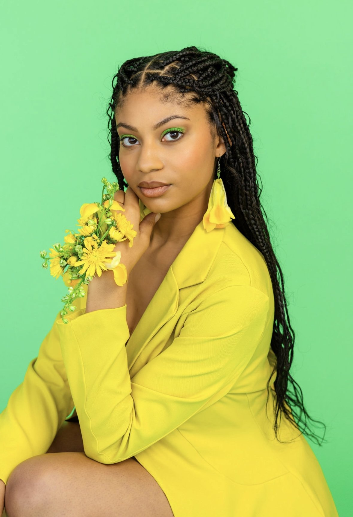 A woman with long, braided hair wearing a yellow blazer and matching yellow earrings, holding a small bouquet of yellow flowers, sitting against a green background.