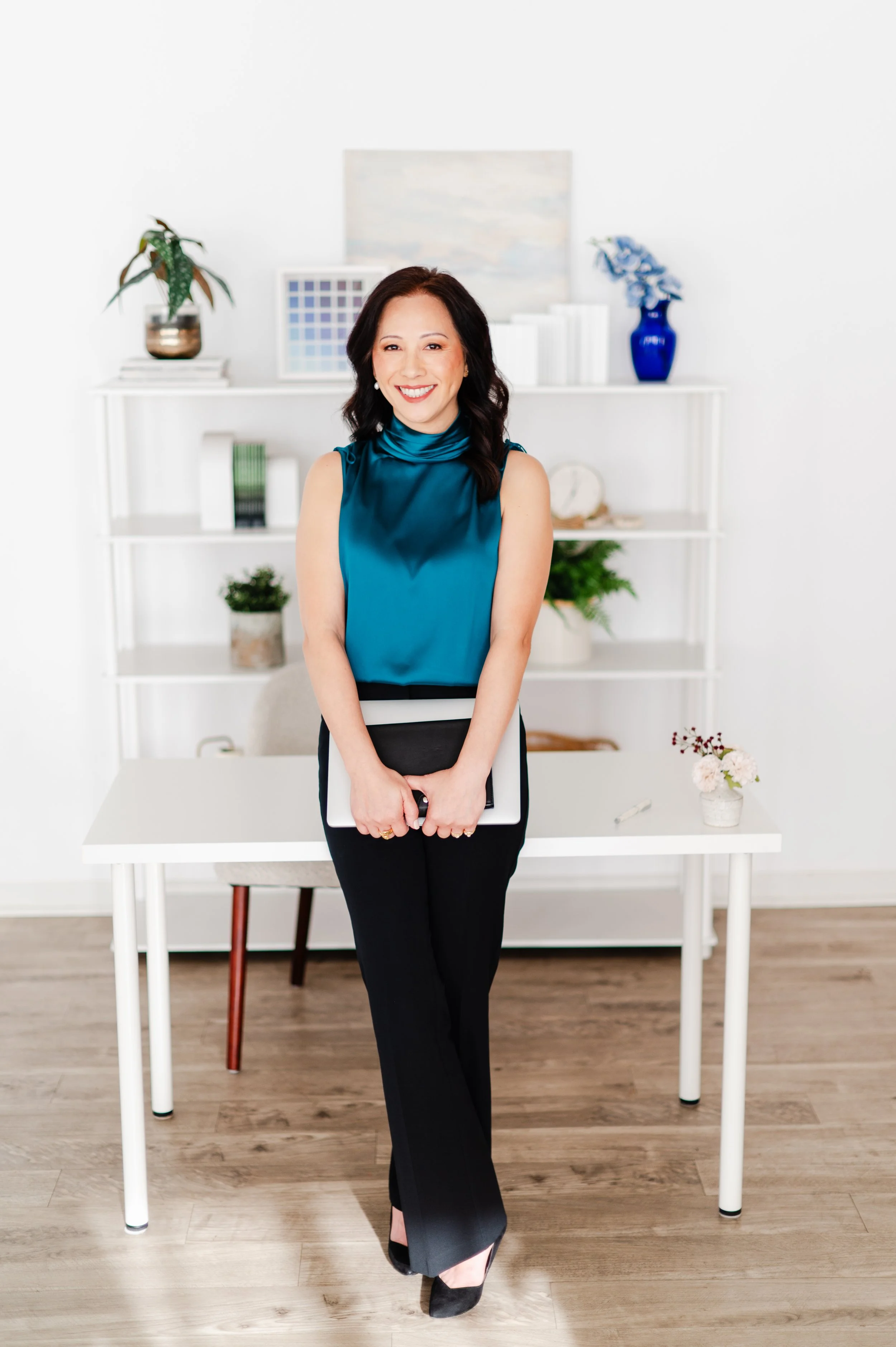 A woman with dark hair in a sleeveless teal satin top and black pants stands in an office with white walls, holding a folder, smiling at the camera.