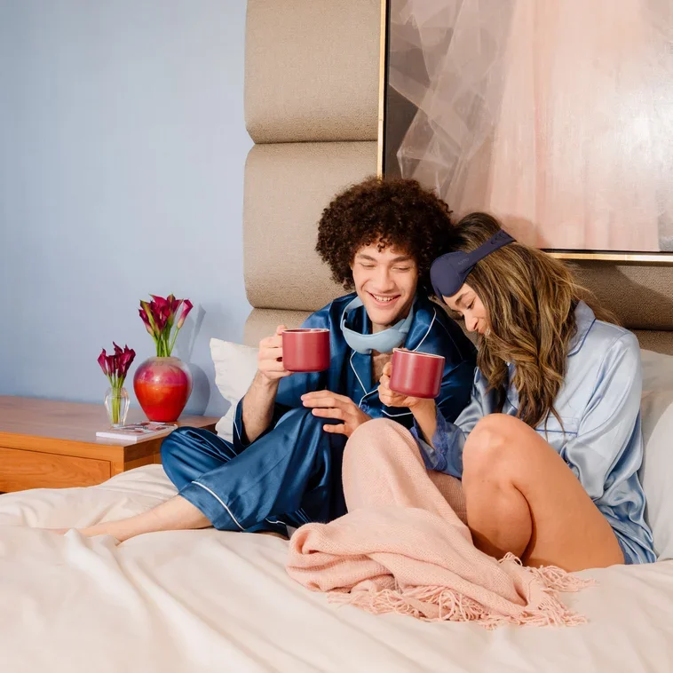 A young couple sitting on a bed, enjoying drinks and breakfast in bed together, smiling and cuddling.