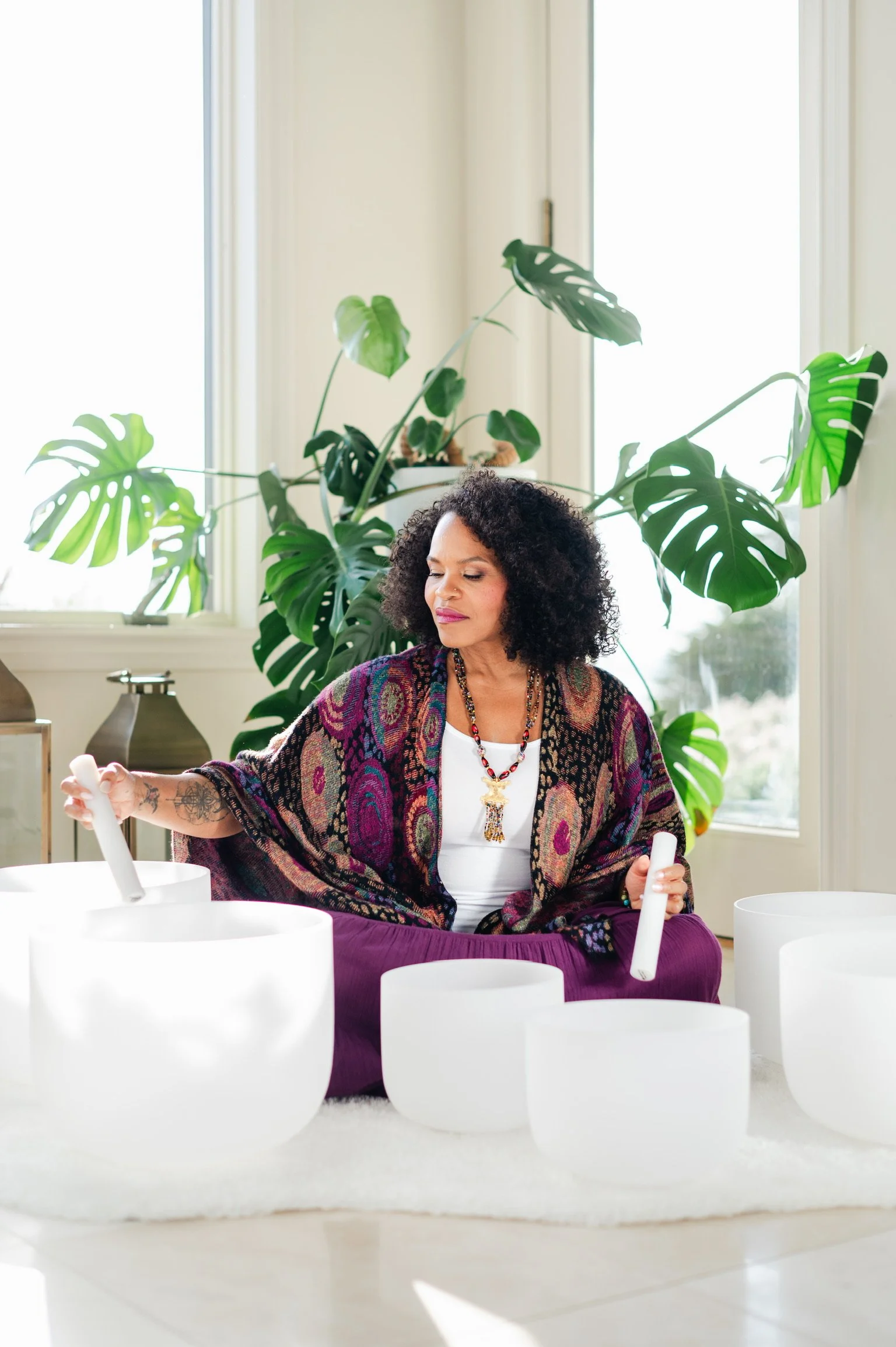 A woman with curly black hair, wearing a colorful shawl and purple pants, playing crystal singing bowls in a bright room with large windows, white walls, and a large monstera plant in the background.