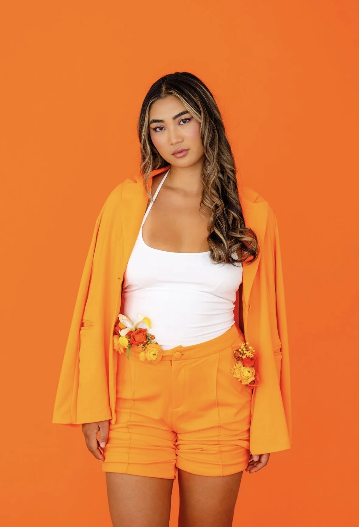 A woman wearing orange shorts and a matching orange jacket over a white top standing against an orange background.