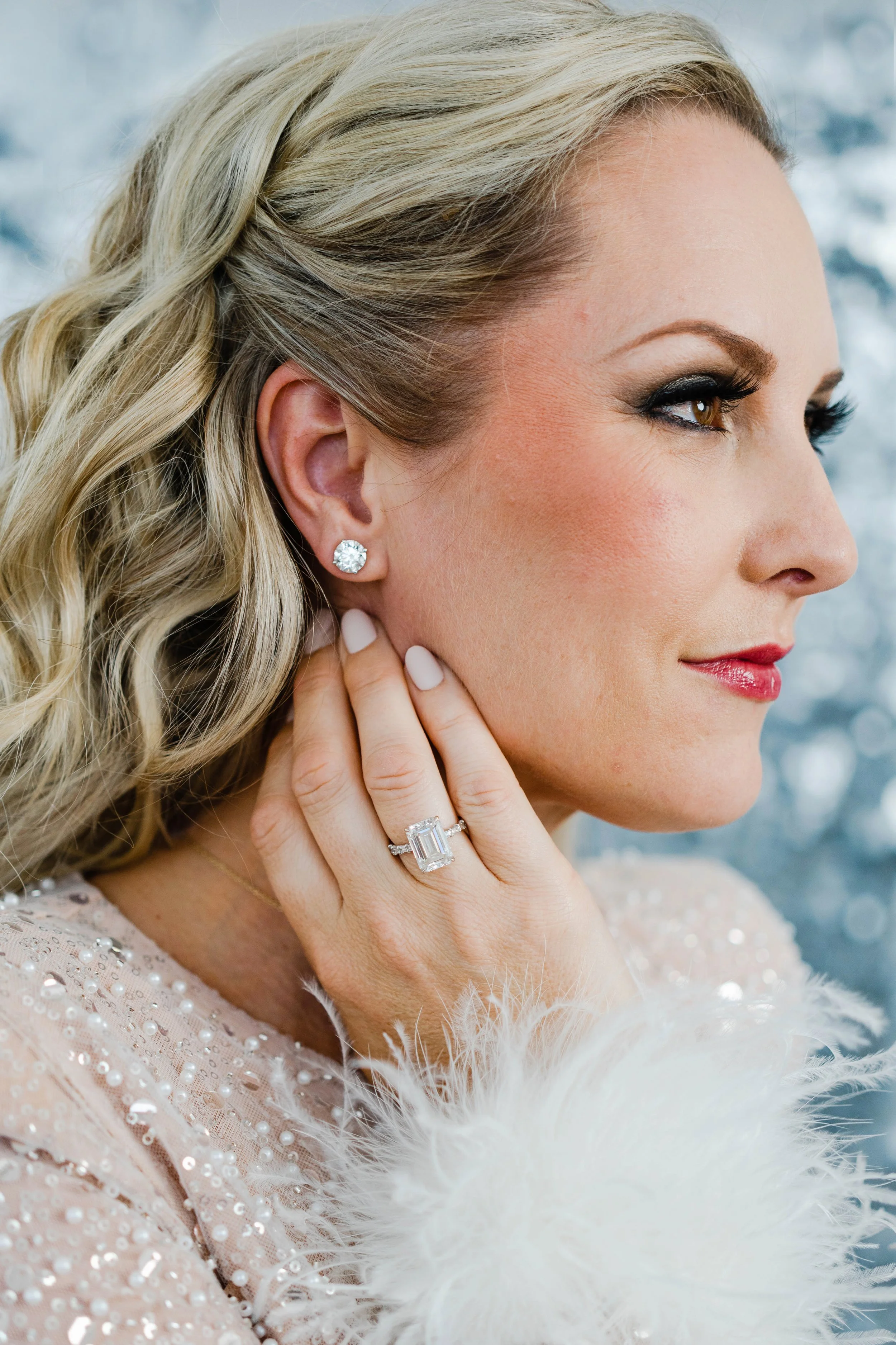 Close-up of a woman with blonde hair, wearing diamond earrings and a large diamond ring, touching her face. She is dressed in a sparkly, light-colored garment with feather details.