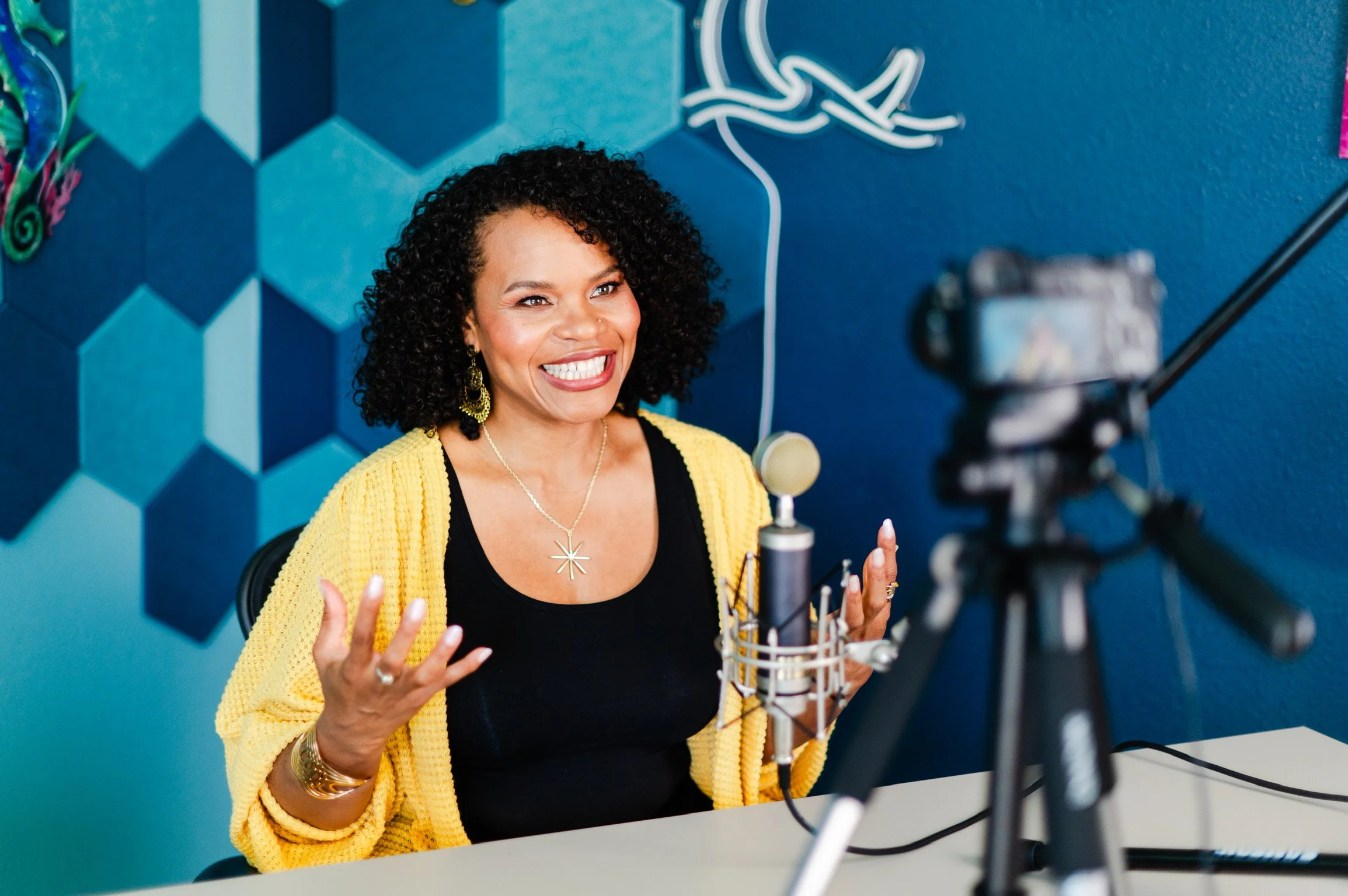 Woman recording a podcast or video with a professional microphone, sitting at a table with a camera on a tripod capturing her.
