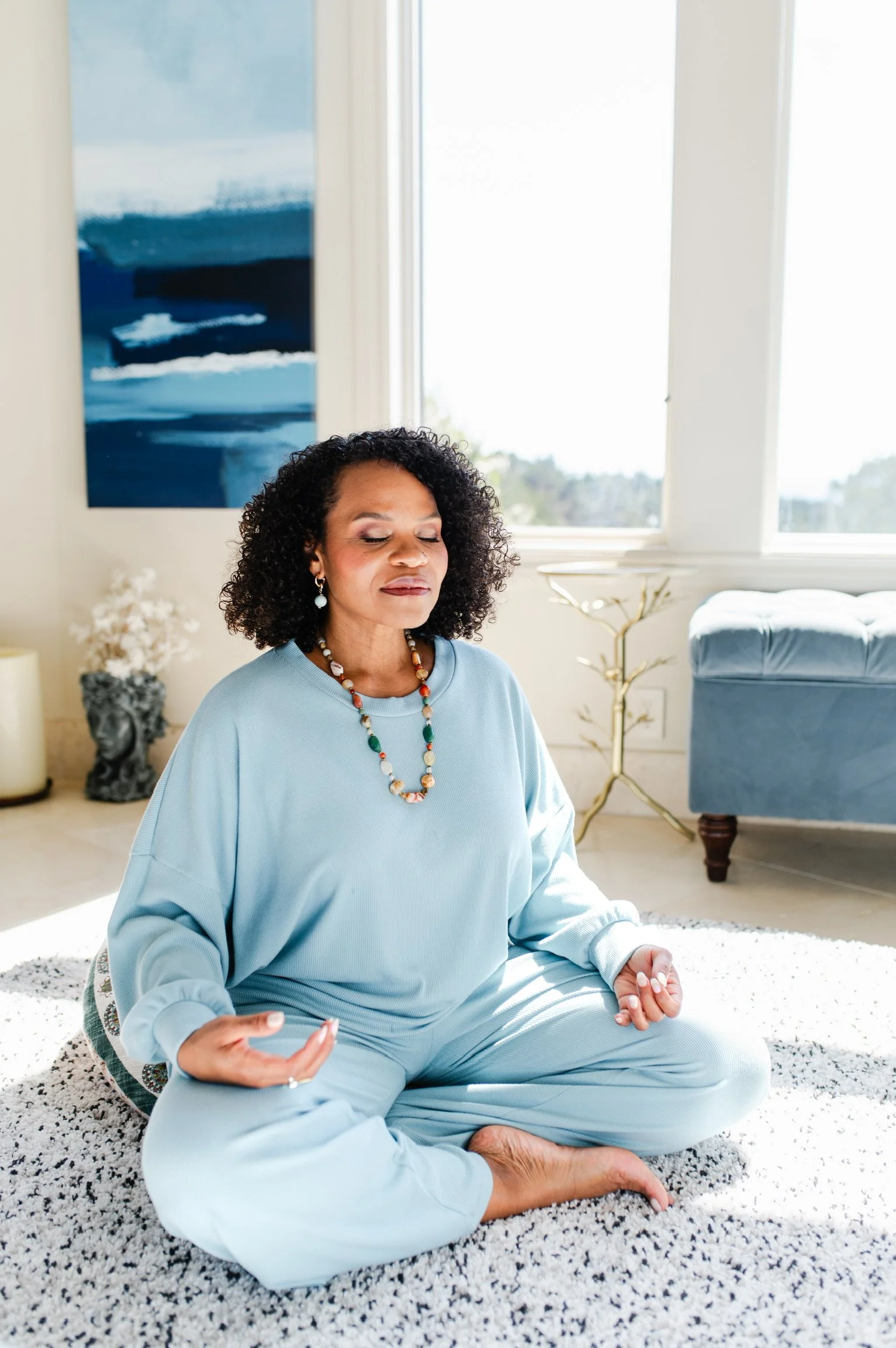 A woman with curly black hair practicing meditation in a bright living room, sitting cross-legged on a white and black speckled rug with eyes closed and hands resting on her knees in a mudra gesture.