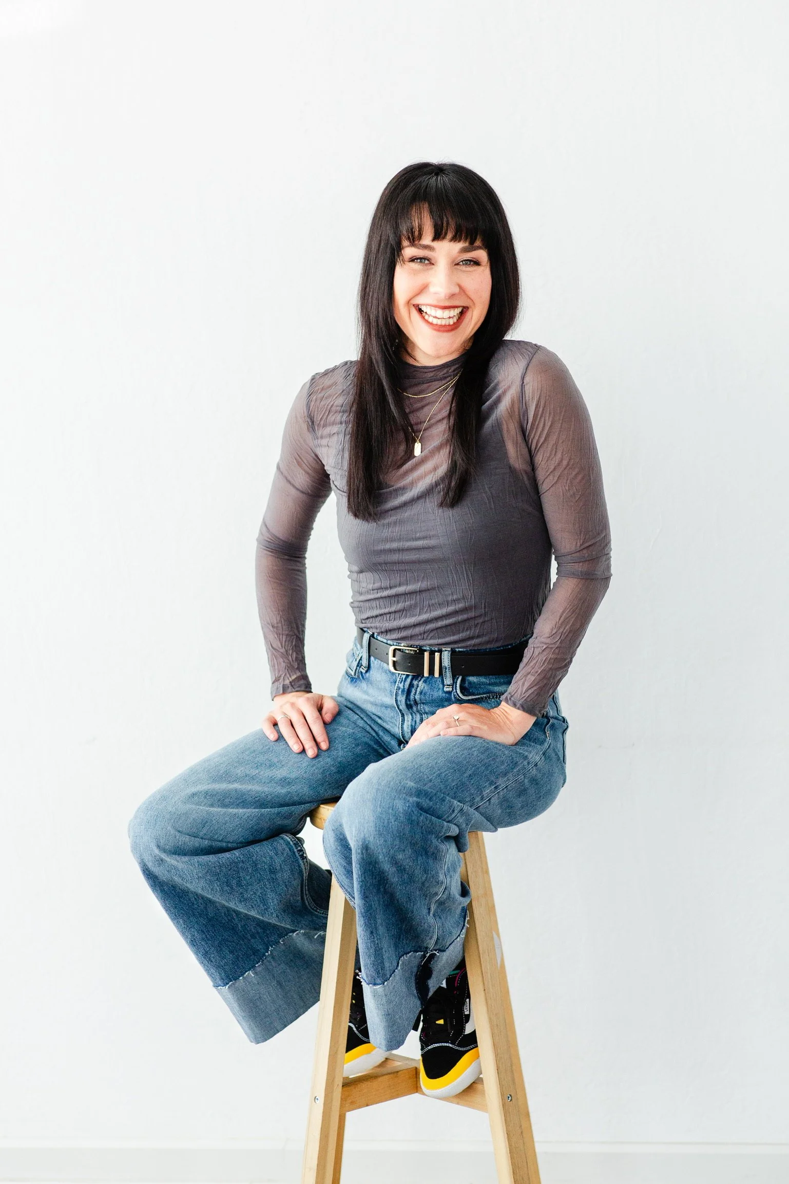 A woman with black hair and bangs smiling while sitting on a wooden stool against a plain white background, wearing a sheer gray long-sleeve top, blue jeans, and black sneakers with yellow accents.