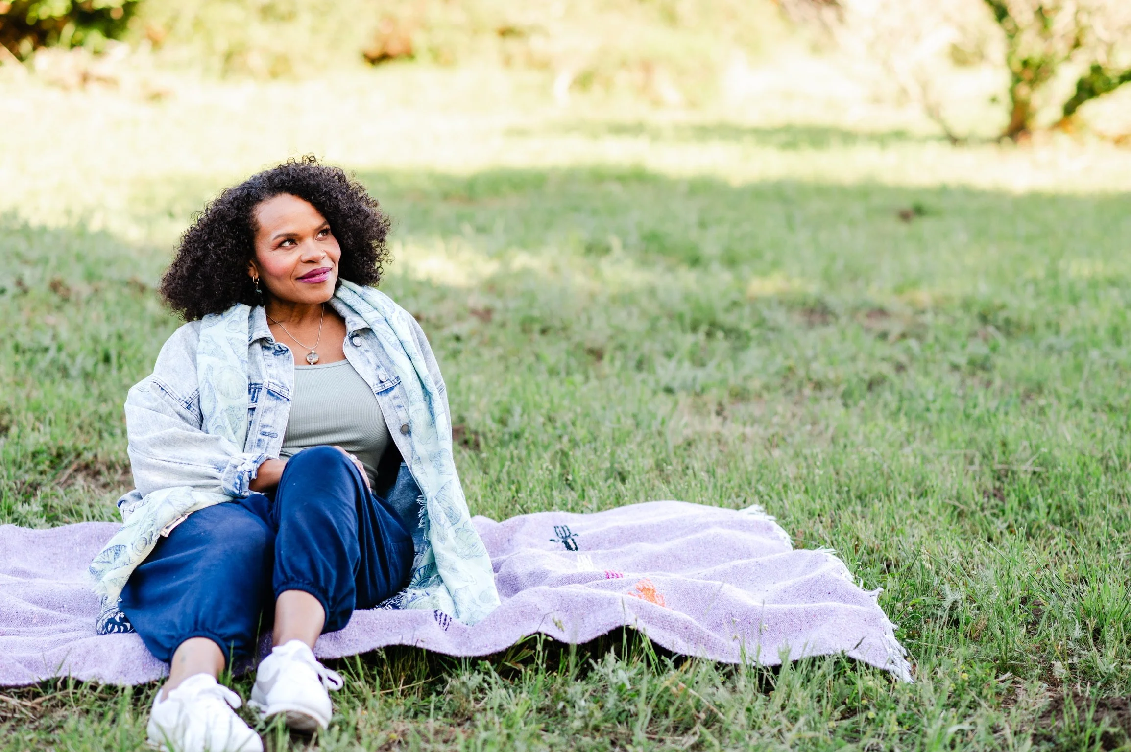 Woman sitting on a purple blanket on the grass in a park, with trees and sunlight in the background.
