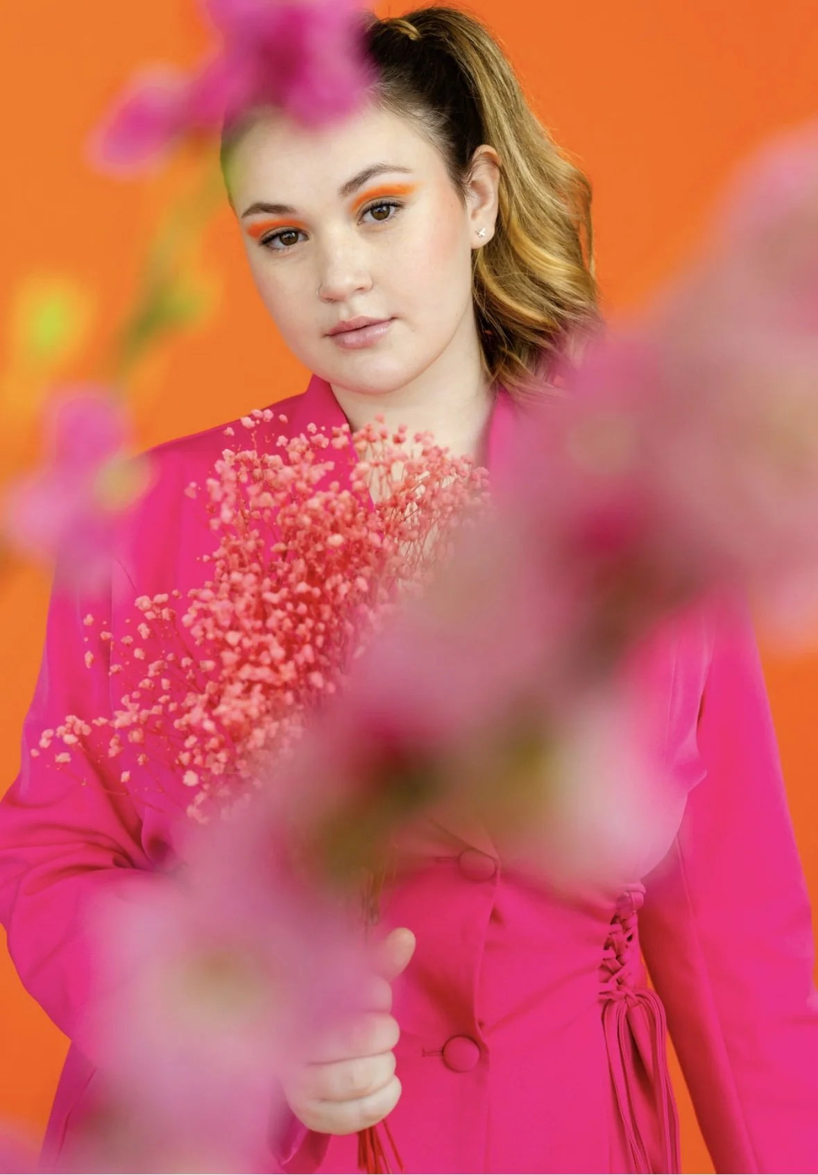 A young woman with wavy blonde hair wearing a pink blazer with button details, looking at the camera against an orange background, surrounded by pink flowers.