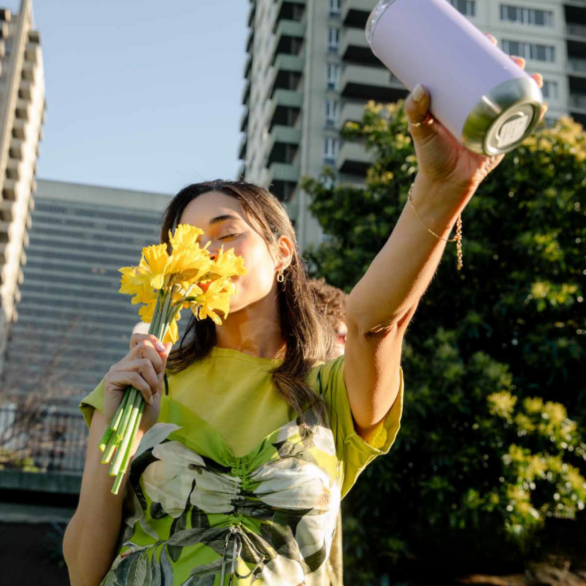 Young woman holding yellow flowers close to her face and taking a selfie outdoors with tall buildings and green trees in the background.