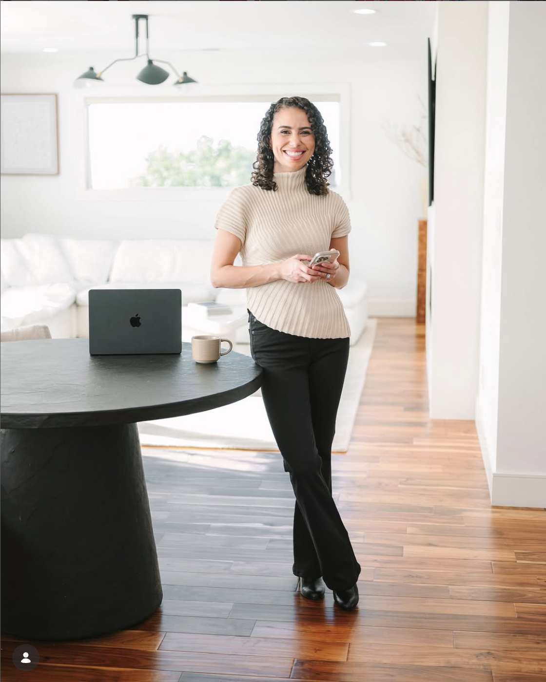 A woman standing in a modern, bright living room with a laptop and coffee mug on a round black table, smiling at the camera while holding a smartphone.