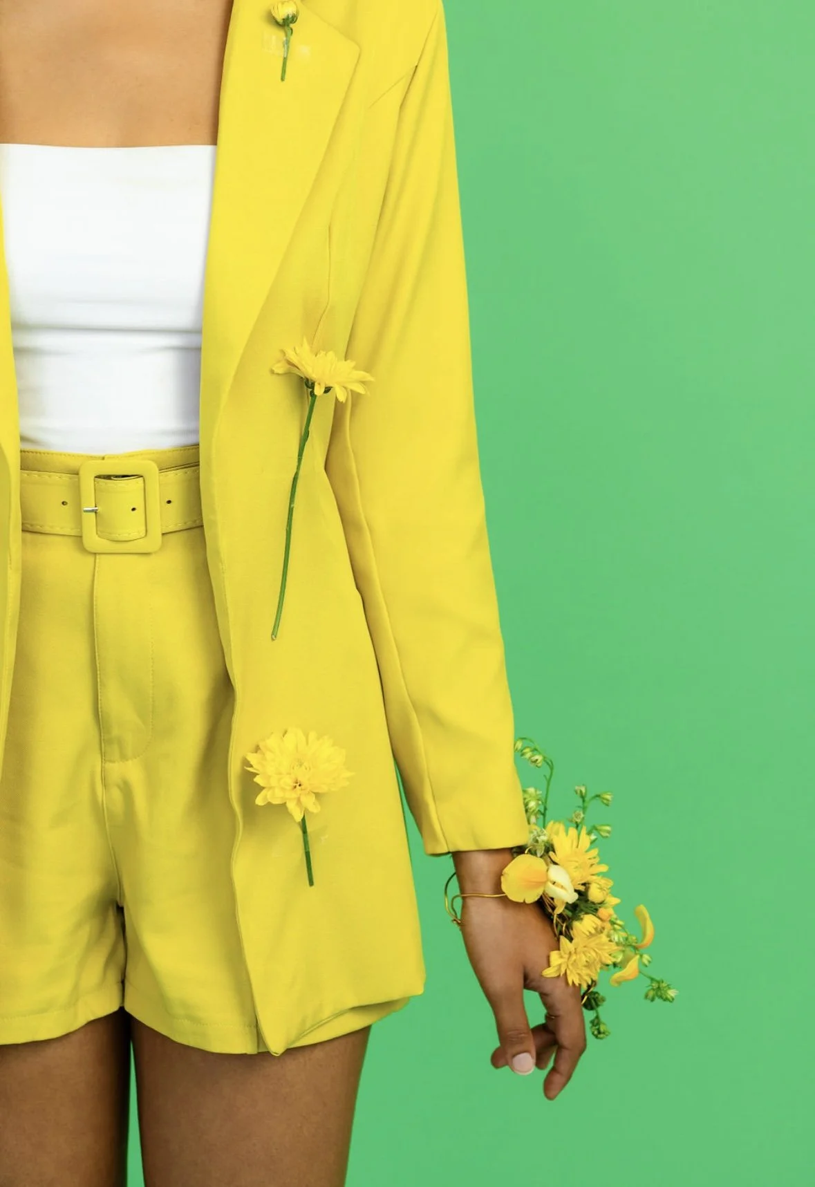 Person wearing a bright yellow blazer, belt, and shorts, with yellow flowers on their blazer, hand, and wrist, standing against a turquoise background.