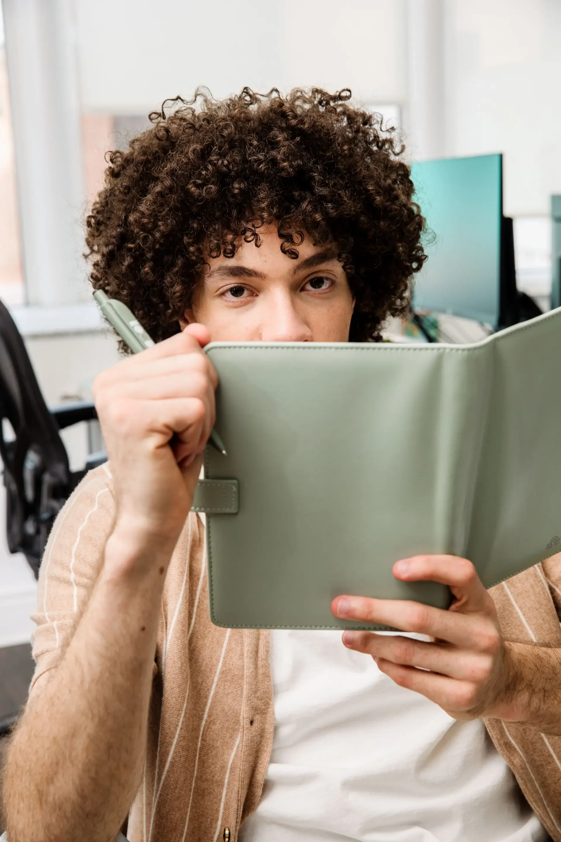 A person with curly hair holding a green tablet partially covering their face while looking at the camera in an office setting.