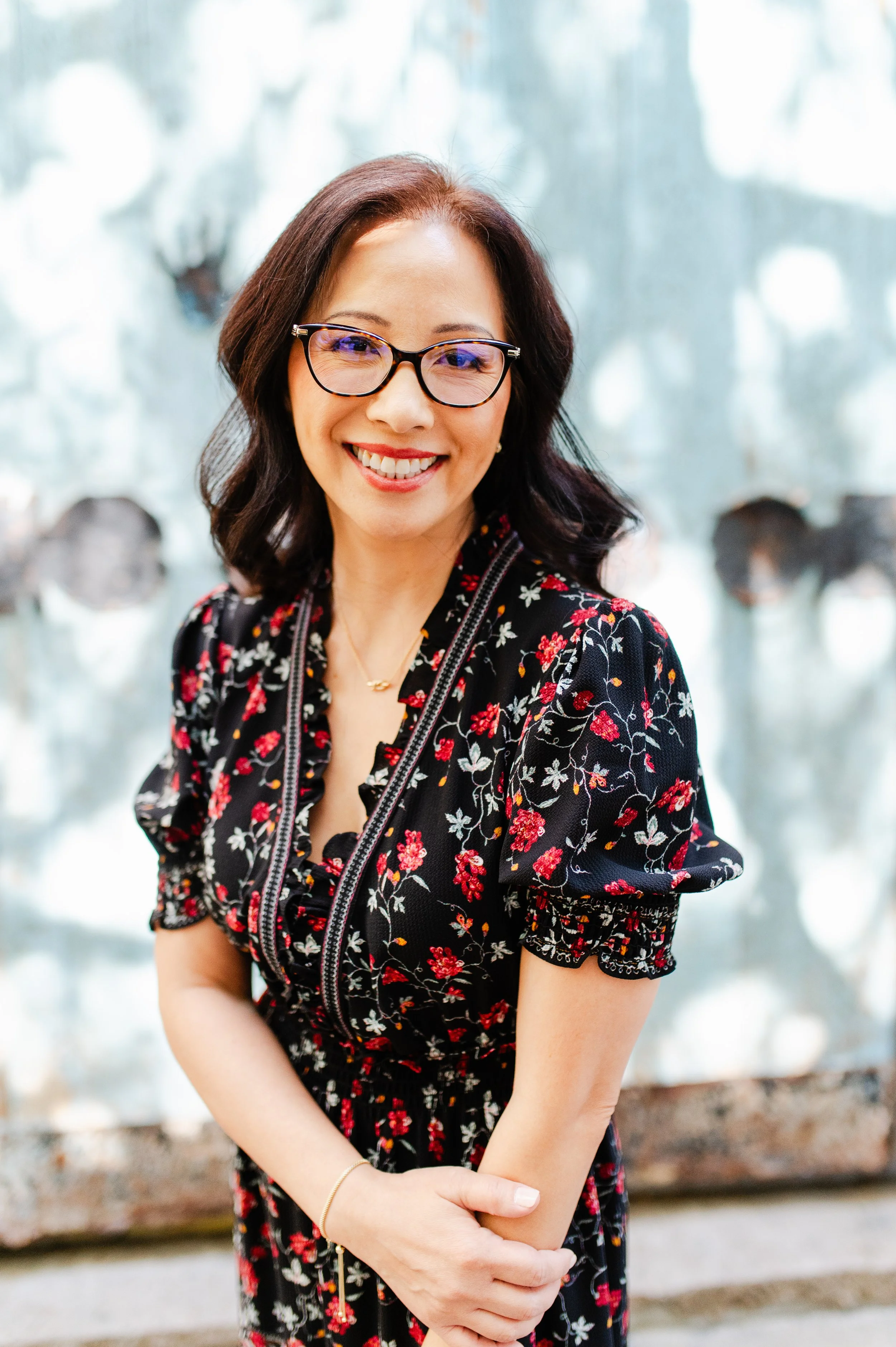 A woman with shoulder-length dark hair, wearing glasses, a black dress with red and white floral pattern, smiling indoors with a blurred background of trees.