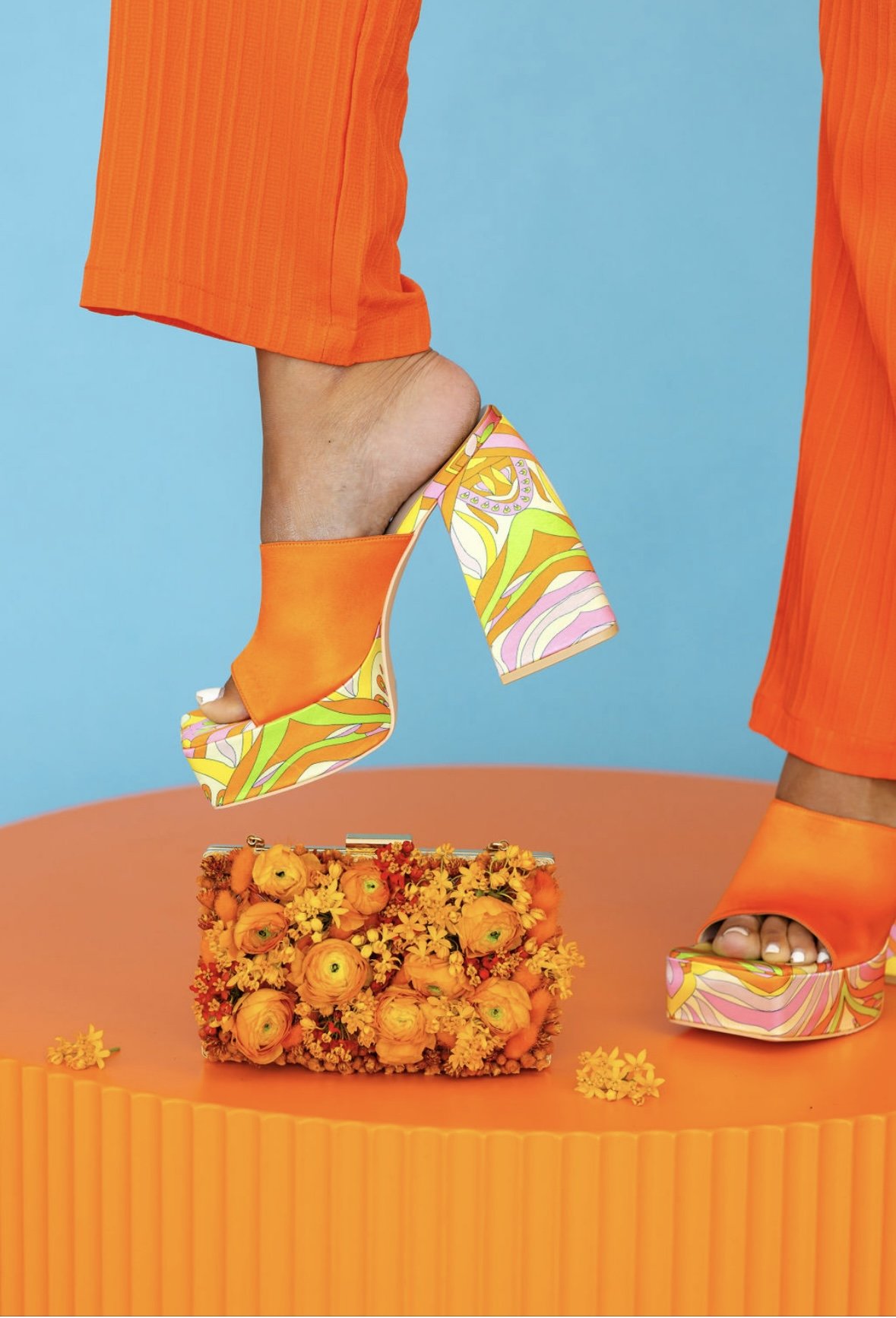 Person wearing orange high-heeled sandals with colorful, abstract patterns, standing on a peach-colored platform with orange flowers around. Person's pants are orange, and they hold a floral handbag with yellow and orange flowers.