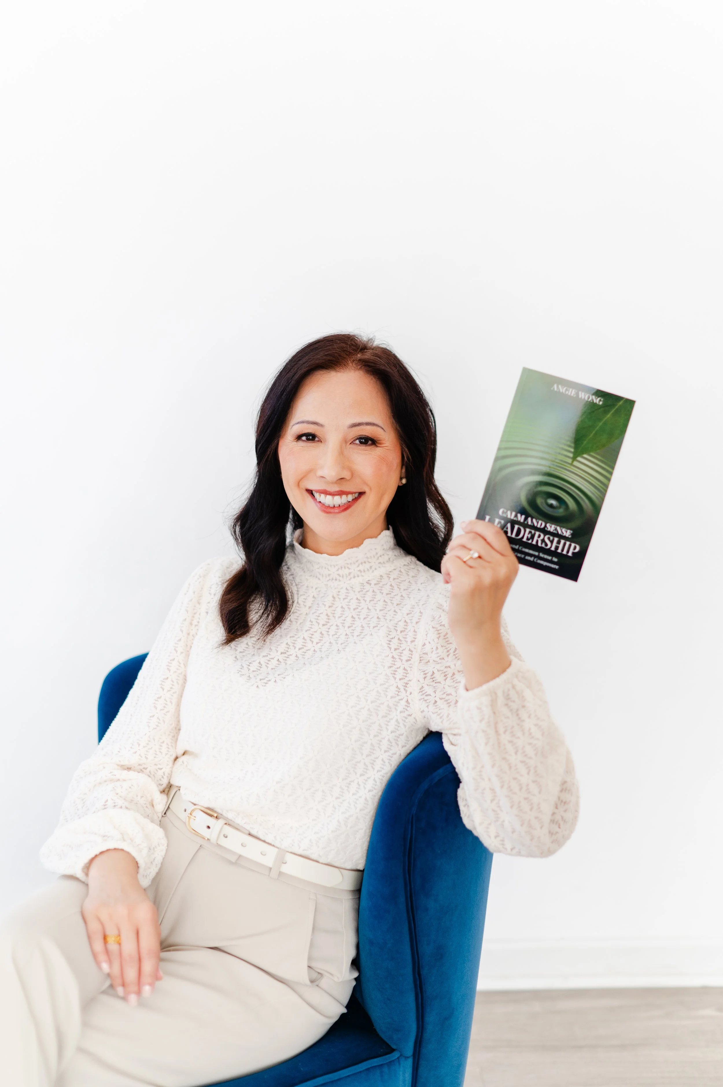 A woman with dark hair, wearing a white textured sweater and beige pants, sitting on a blue chair against a white background. She is smiling and holding a book titled 'Calm and Sense Leadership' by Angie Wong in her right hand.
