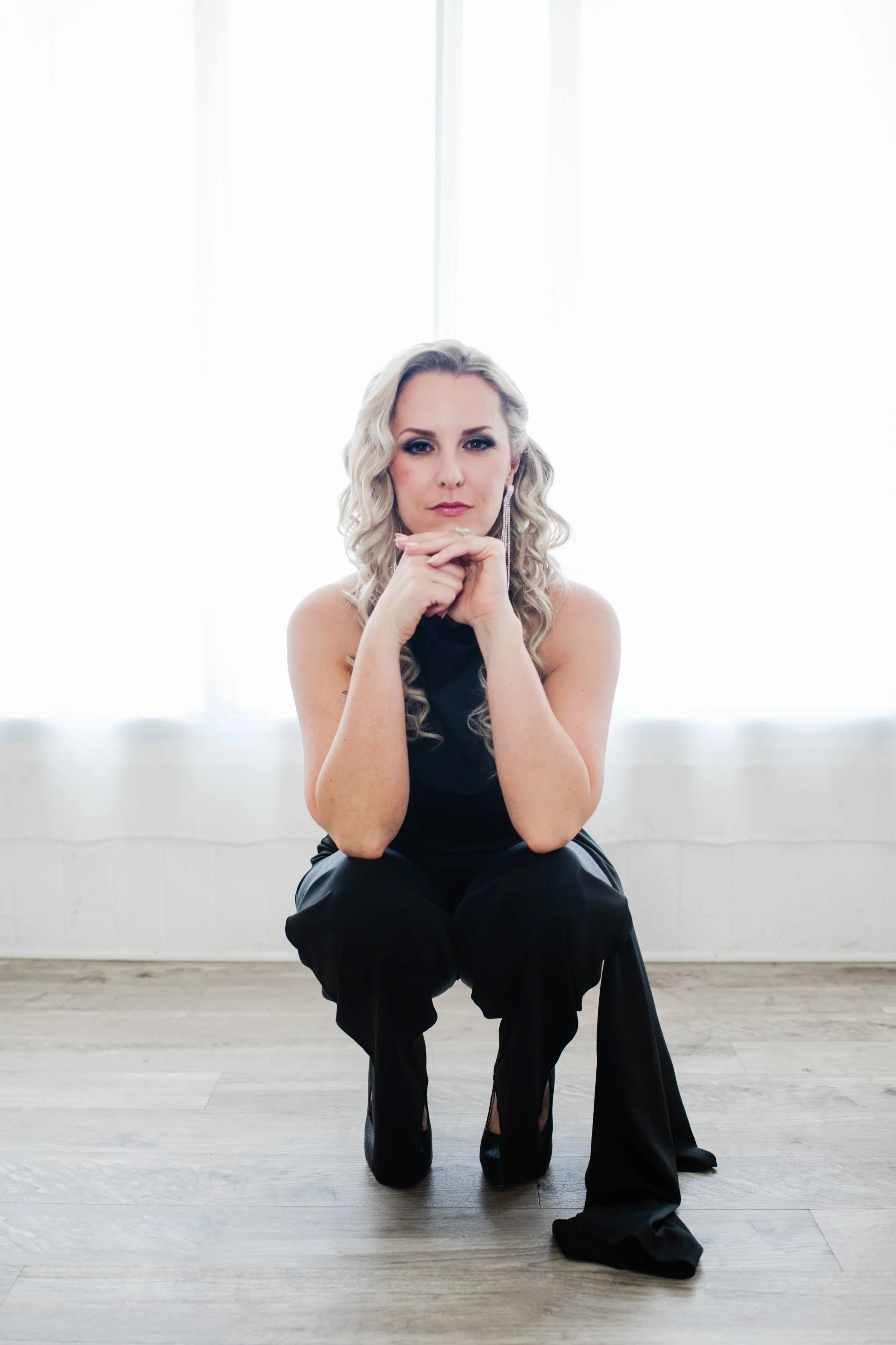 Woman with blonde curly hair in a black outfit, squatting on a wooden floor in front of bright backlit windows.