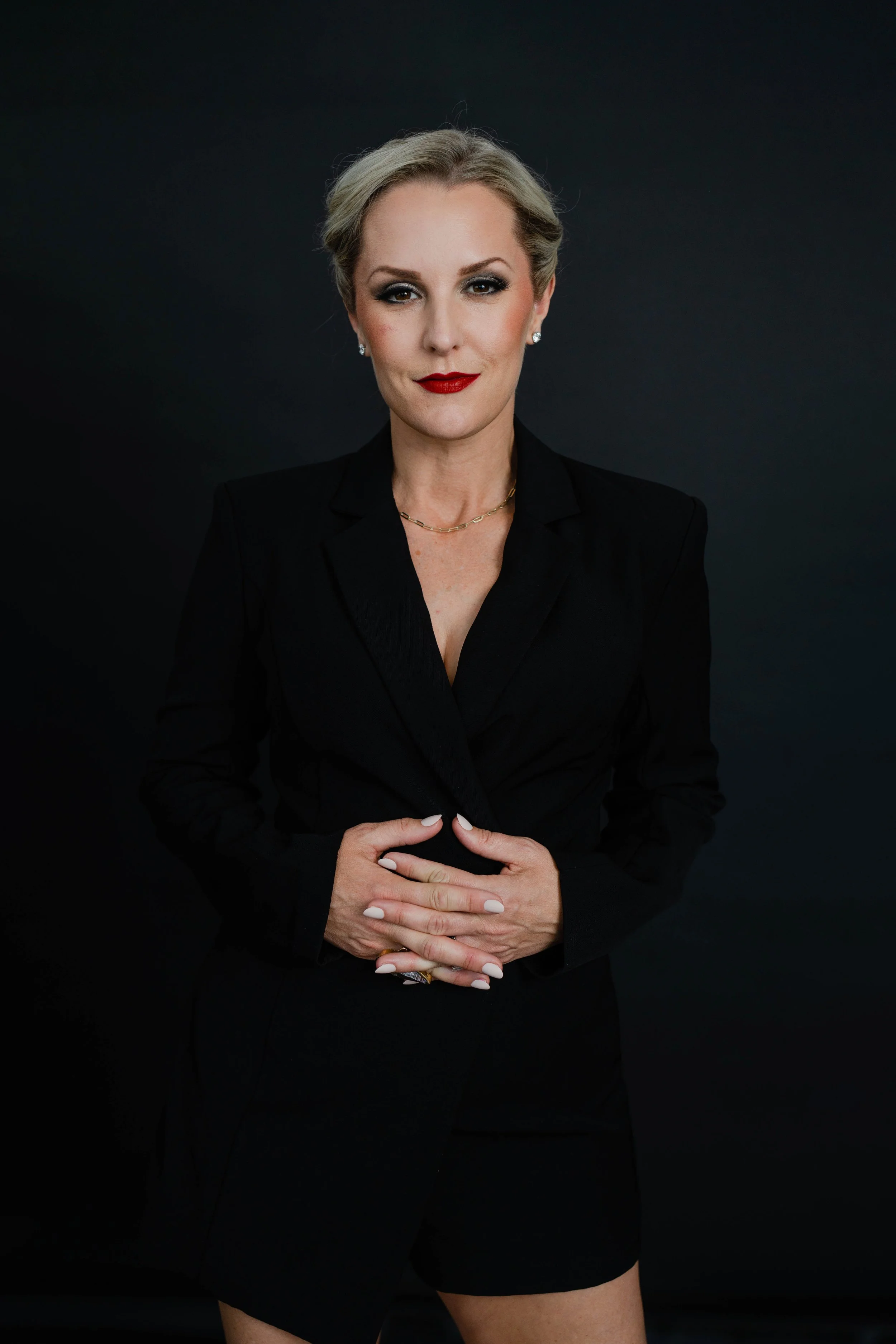 A confident woman with blonde hair styled back, wearing a black blazer and shorts, posing against a plain dark background