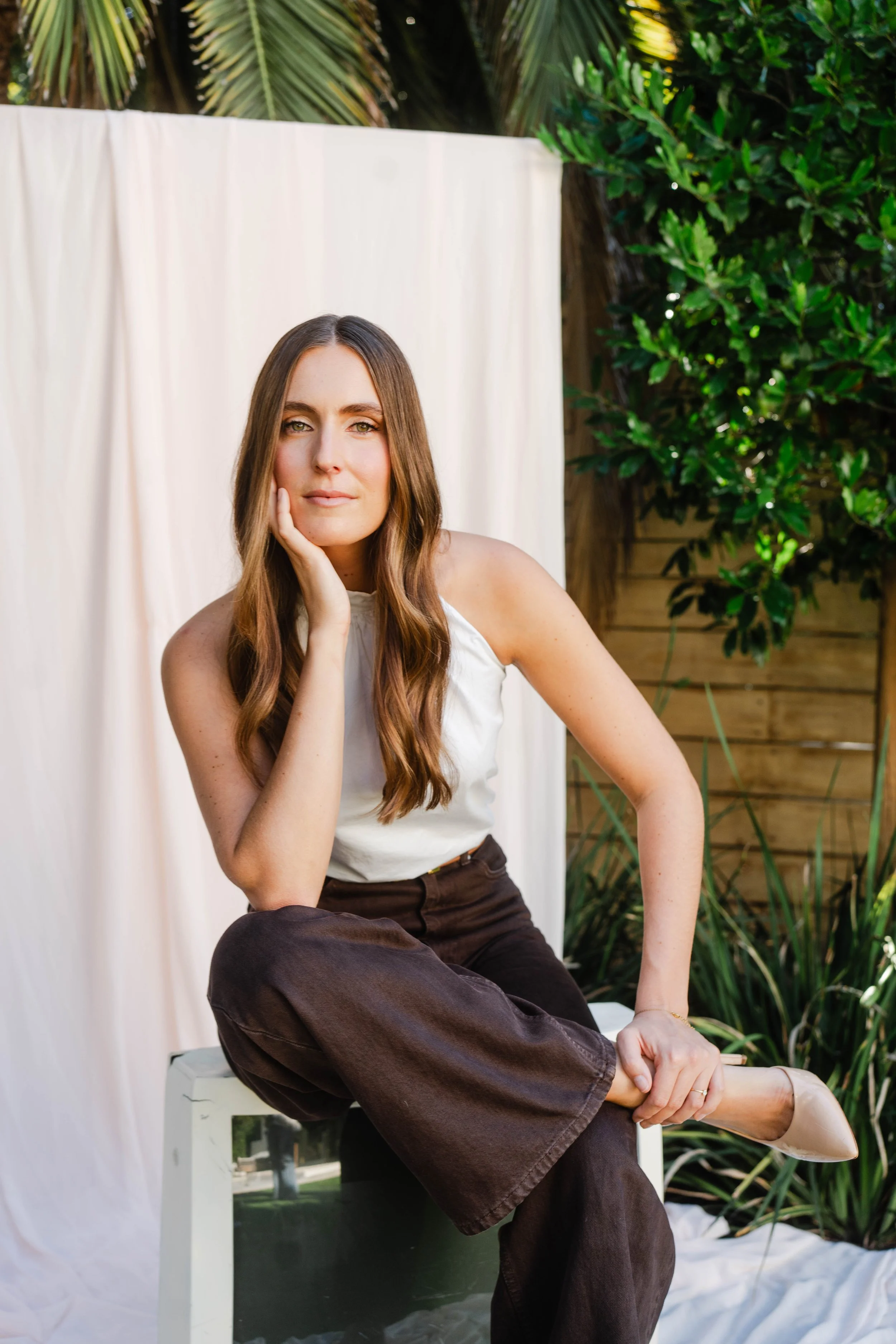 A woman with long brown hair, wearing a white sleeveless top and brown pants, sitting outdoors on a small white box, with her left knee raised and her right hand resting on her chin, in front of a white cloth backdrop and greenery.