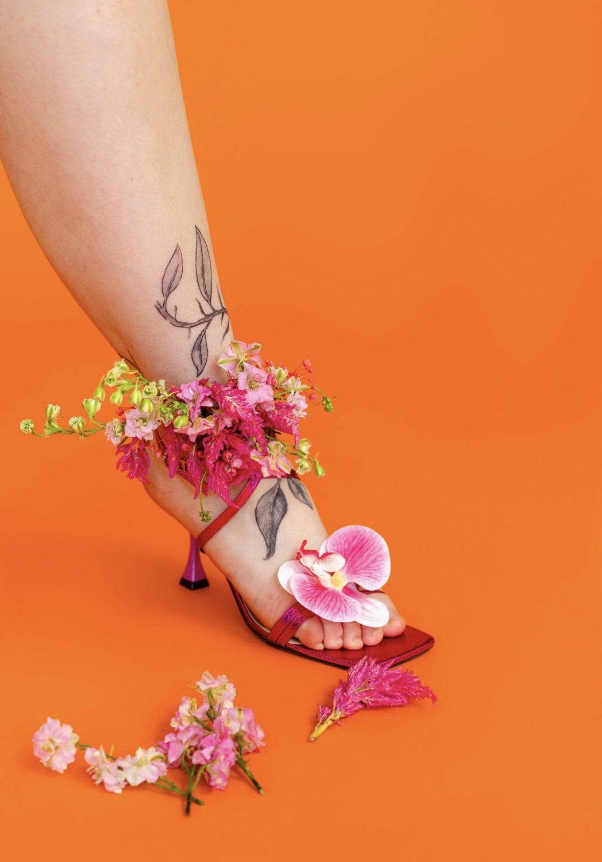 A woman is standing on an orange surface, wearing a red high-heeled sandal decorated with pink flowers. Her leg has a tattoo of leaves and a slender branch. There are pink and white orchids and other flowers around her ankle and foot, with one orchid