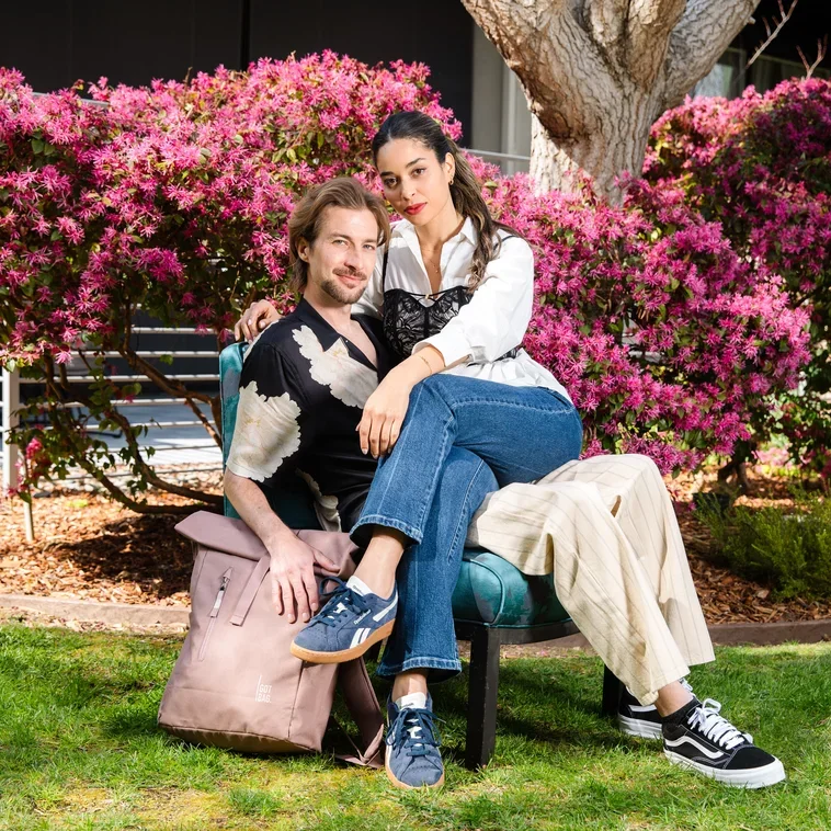 Two young adults sitting on a park bench in front of vibrant pink flowering bushes, with one person sitting on the other's lap, outdoors on a sunny day.
