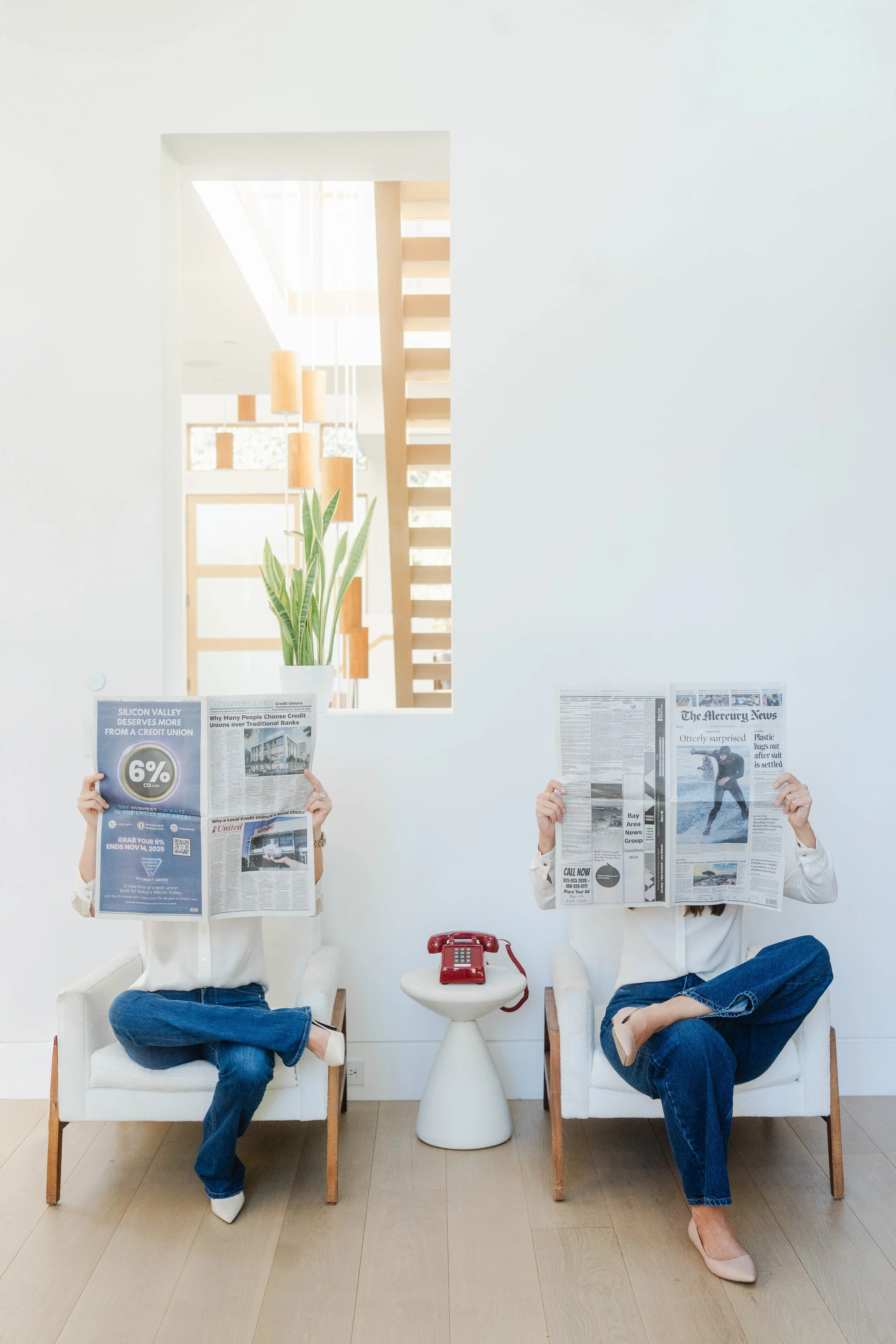 Two women sitting in white armchairs, reading newspapers, with a red telephone on a white side table between them. A plant in a white pot is behind them, and a staircase with wooden steps is visible through a window on the wall.