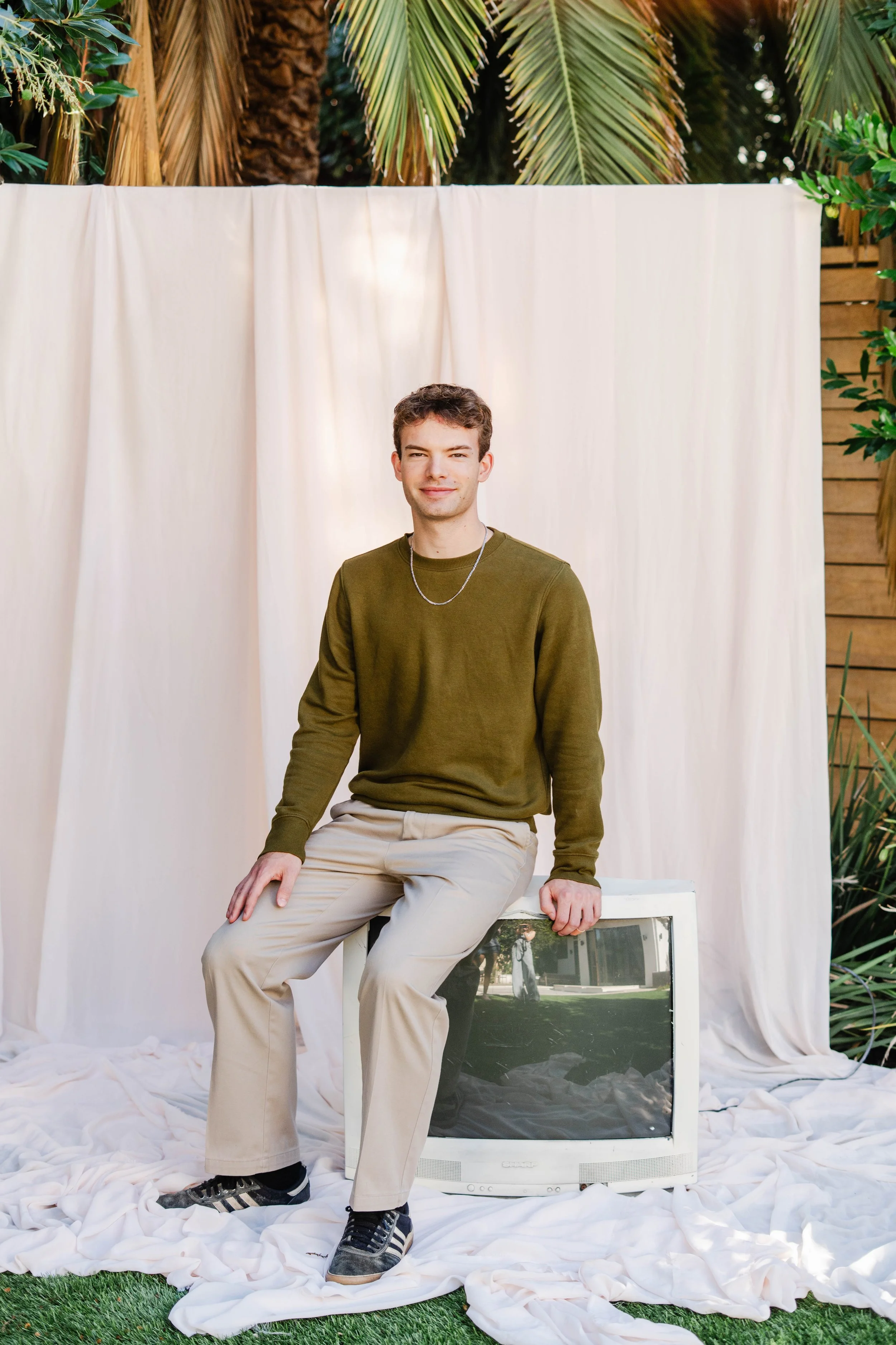 A young man in a green long-sleeve shirt and beige pants sitting on an old television, posing outdoors in front of a white backdrop with tropical plants around.