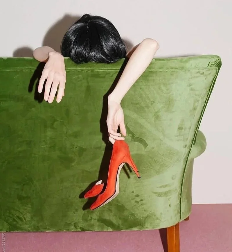A person with black hair is leaning over the back of a green velvet armchair, holding a red high-heeled shoe that hangs down.