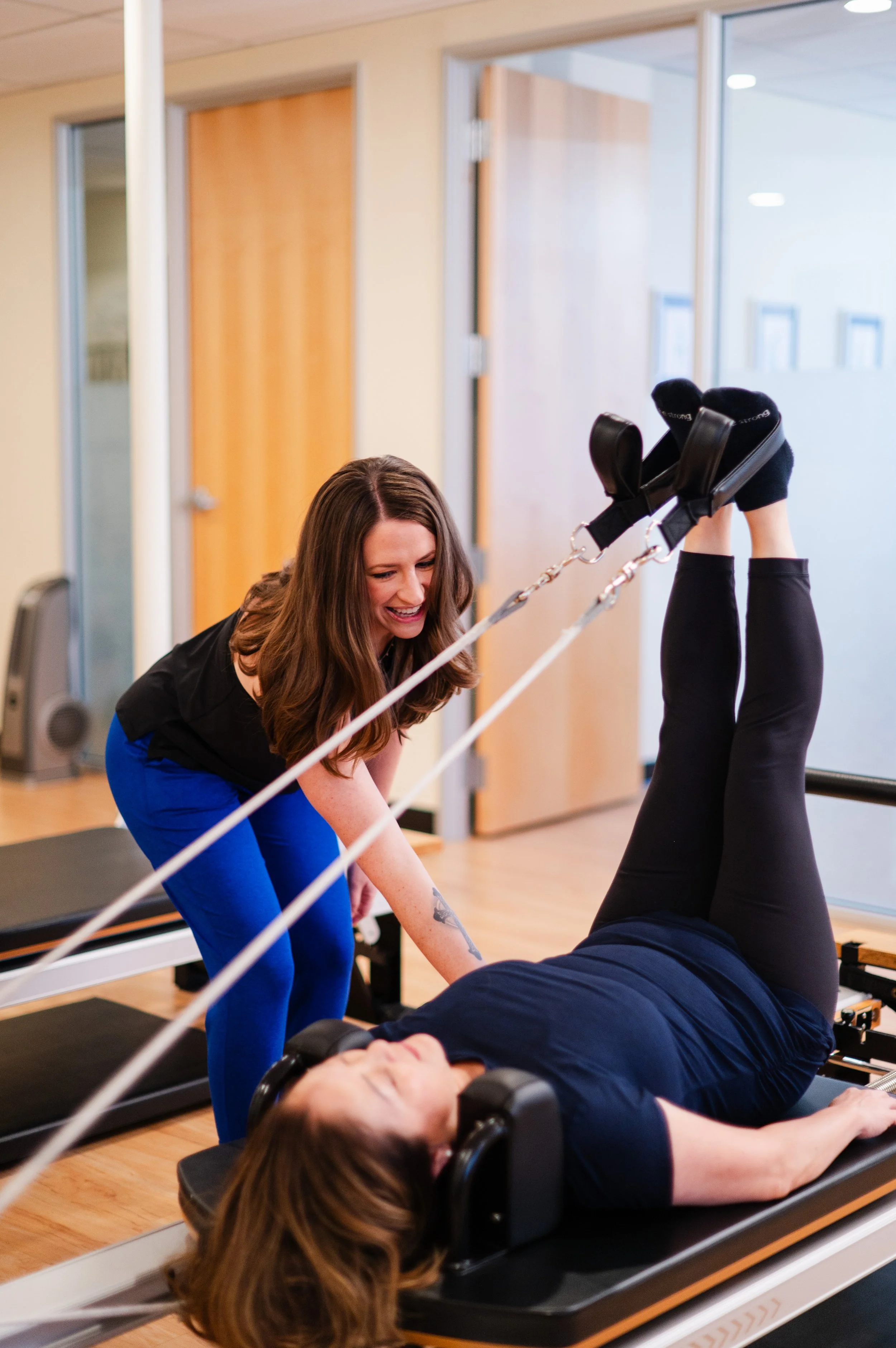 A woman lying on a Pilates reformer machine during a workout, assisted by a trainer in a fitness studio.