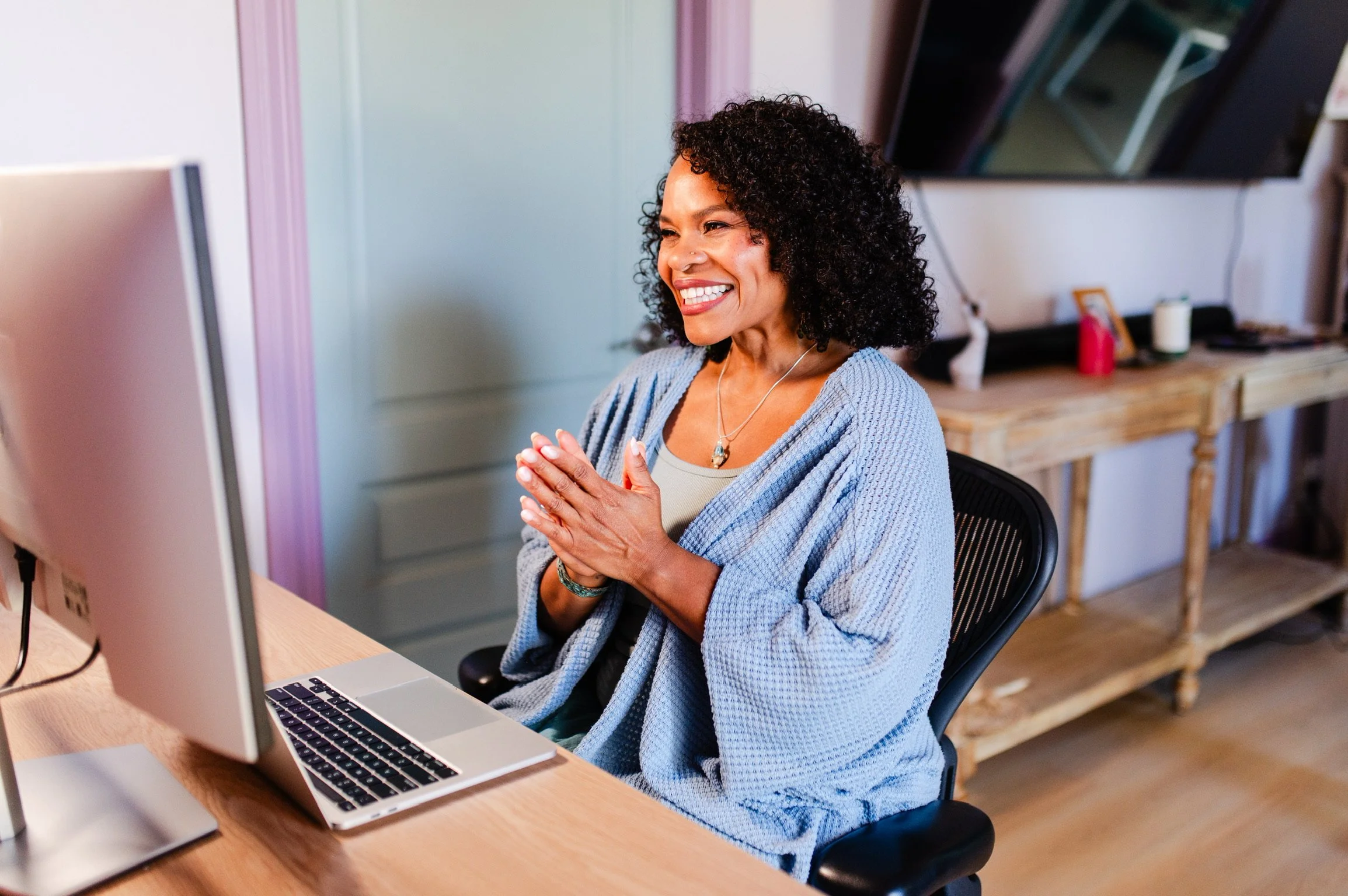A woman with dark curly hair smiling at her computer with hands clasped in front of her chest in a home office