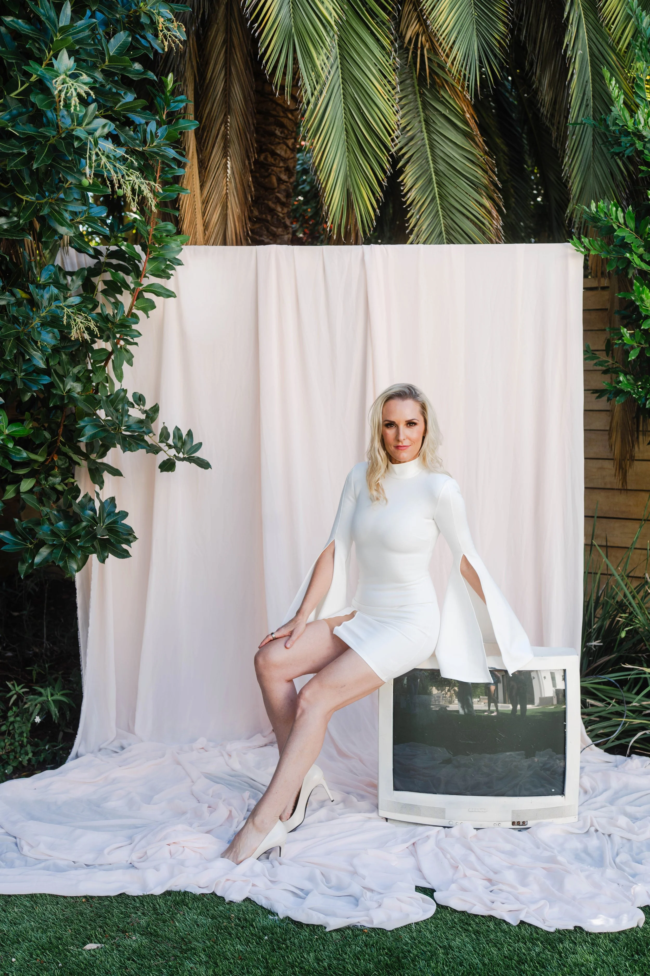 A woman in a white dress and high heels sitting on a chair with an old television underneath her, outdoors with greenery and palm trees in the background.