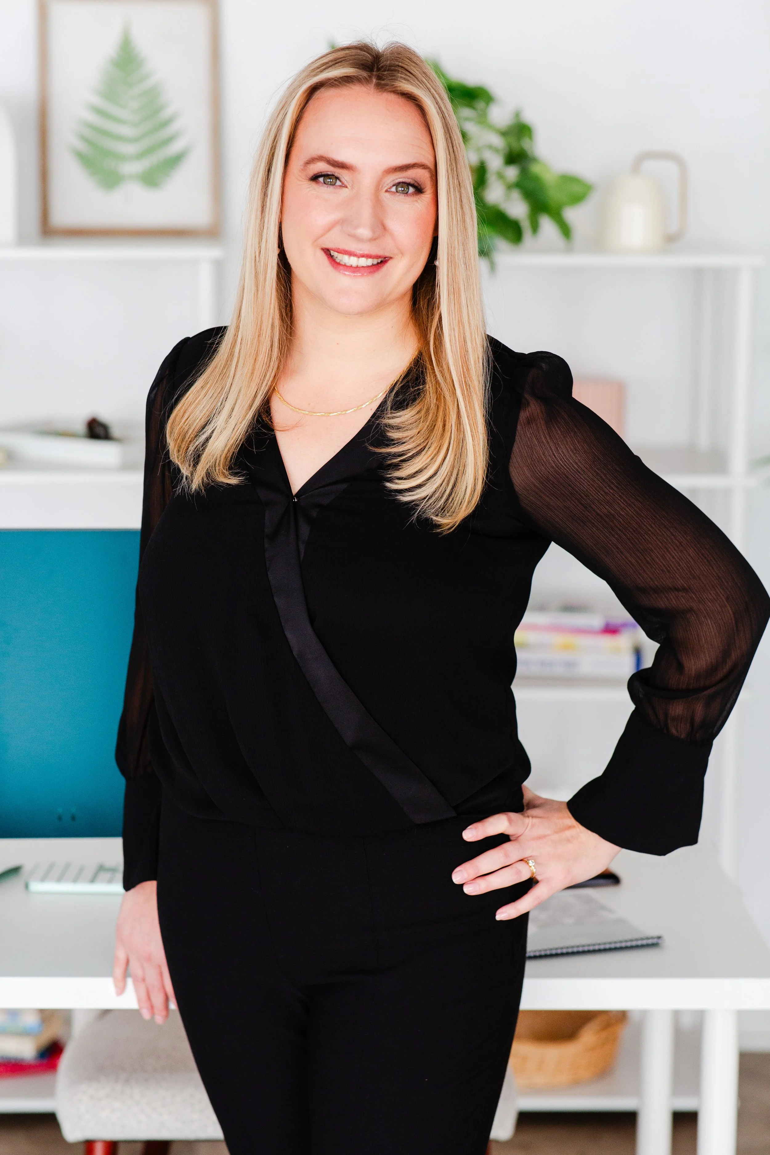 A woman with blonde hair, wearing a black blouse, standing in an office or home office environment, smiling with her hand on her hip.