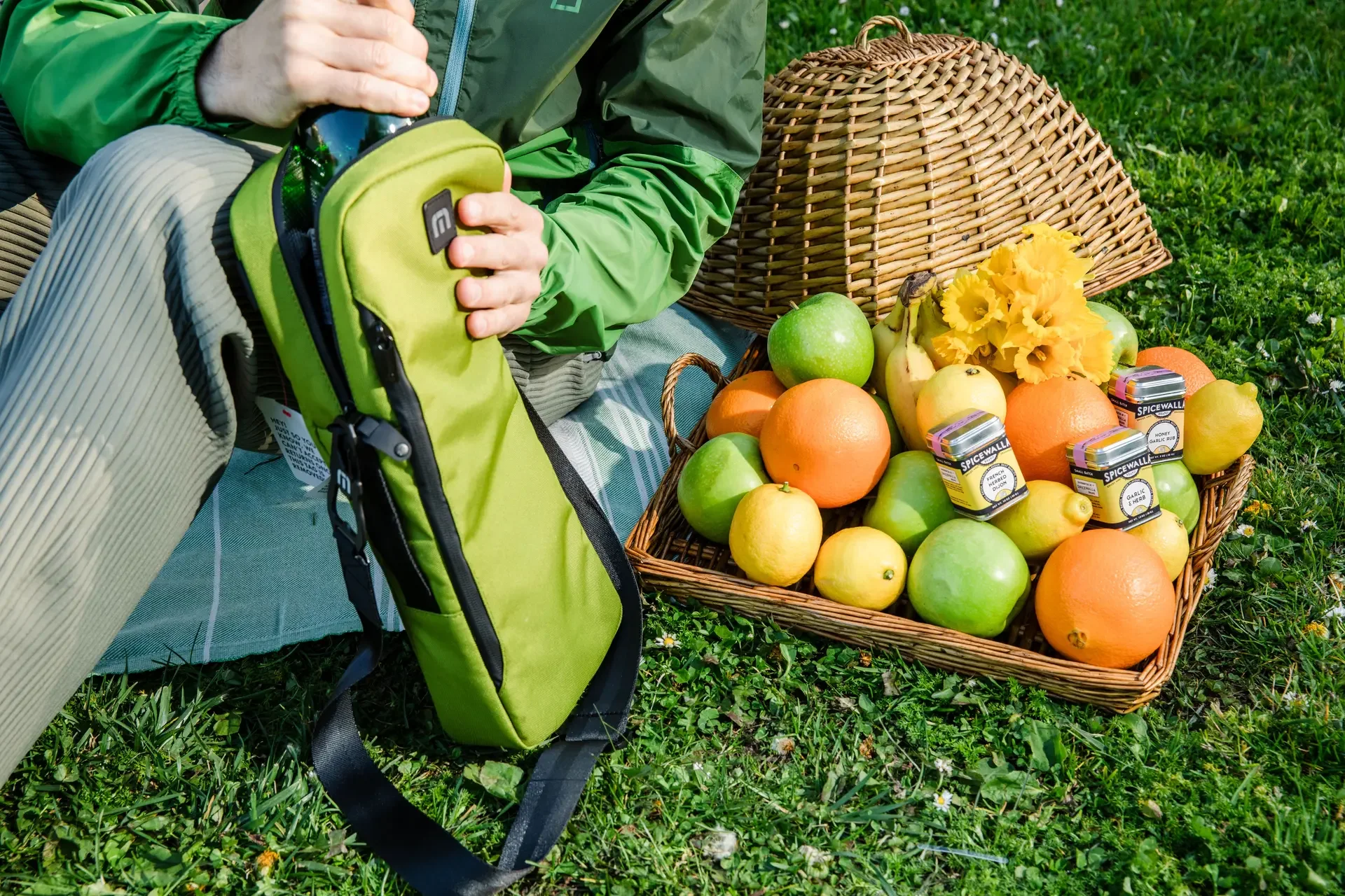 A basket of fresh fruits including green apples, lemons, oranges, and bananas, with a bunch of yellow flowers, placed on grass. Part of a person wearing green and tan pants and holding a green backpack is visible, sitting on a mat outdoors.