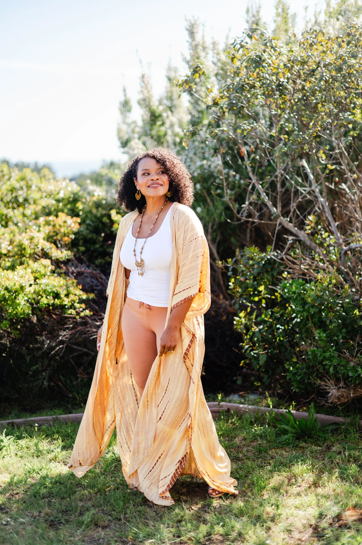 A woman with curly hair, wearing a white top and beige pants, standing outdoors with greenery in the background.
