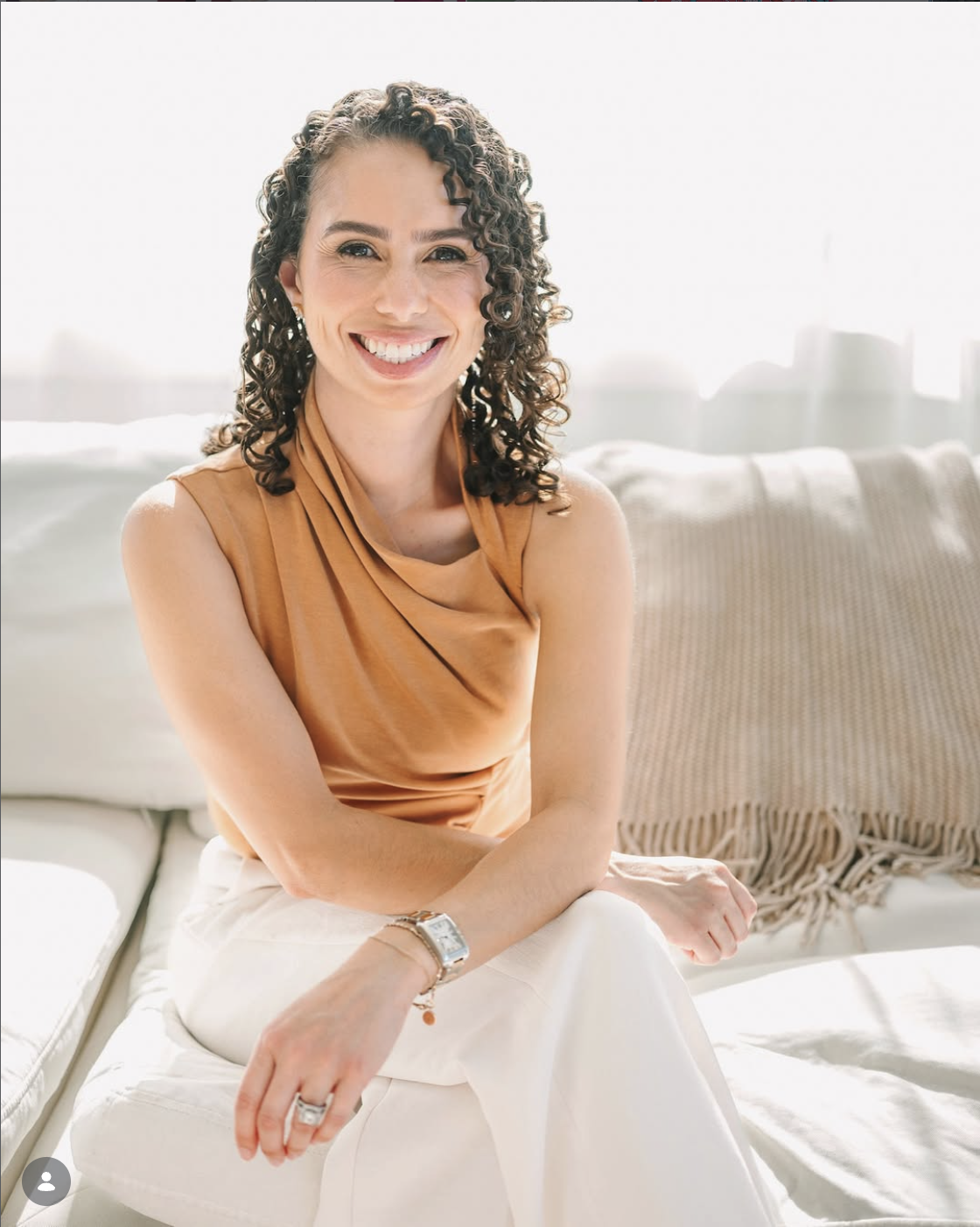 A smiling woman with curly hair wearing a sleeveless bronze top and white pants, sitting on a white couch with a beige cushion and neutral background.